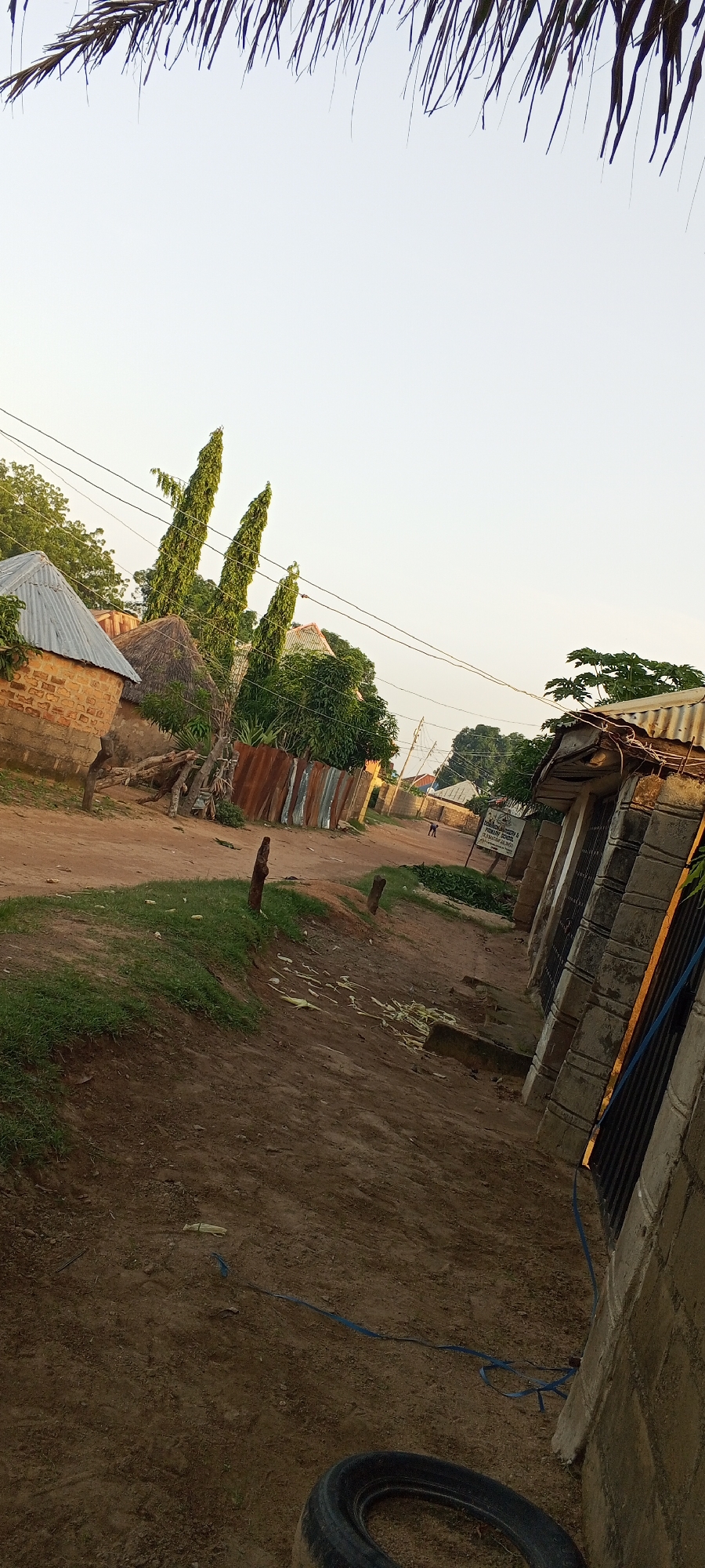 The image depicts a dirt road in a village, possibly in Jalingo, Nigeria. The road is unpaved and appears to be the main thoroughfare of the settlement. On either side of the road are various structures, including homes made of mud brick with corrugated metal roofs and thatched roofs. Some of these structures are partially obscured by trees and vegetation. There are also fences made of corrugated metal and some wooden debris. In the foreground, there is a patch of grass on the left and the dirt road continues to the right. A tire lies on the ground in the bottom right corner. In the distance, a person can be seen walking on the road. There are power lines running overhead, suggesting the presence of electricity. The sky is clear and bright, indicating it is daytime. The overall scene suggests a rural or semi-rural setting with a modest level of development.