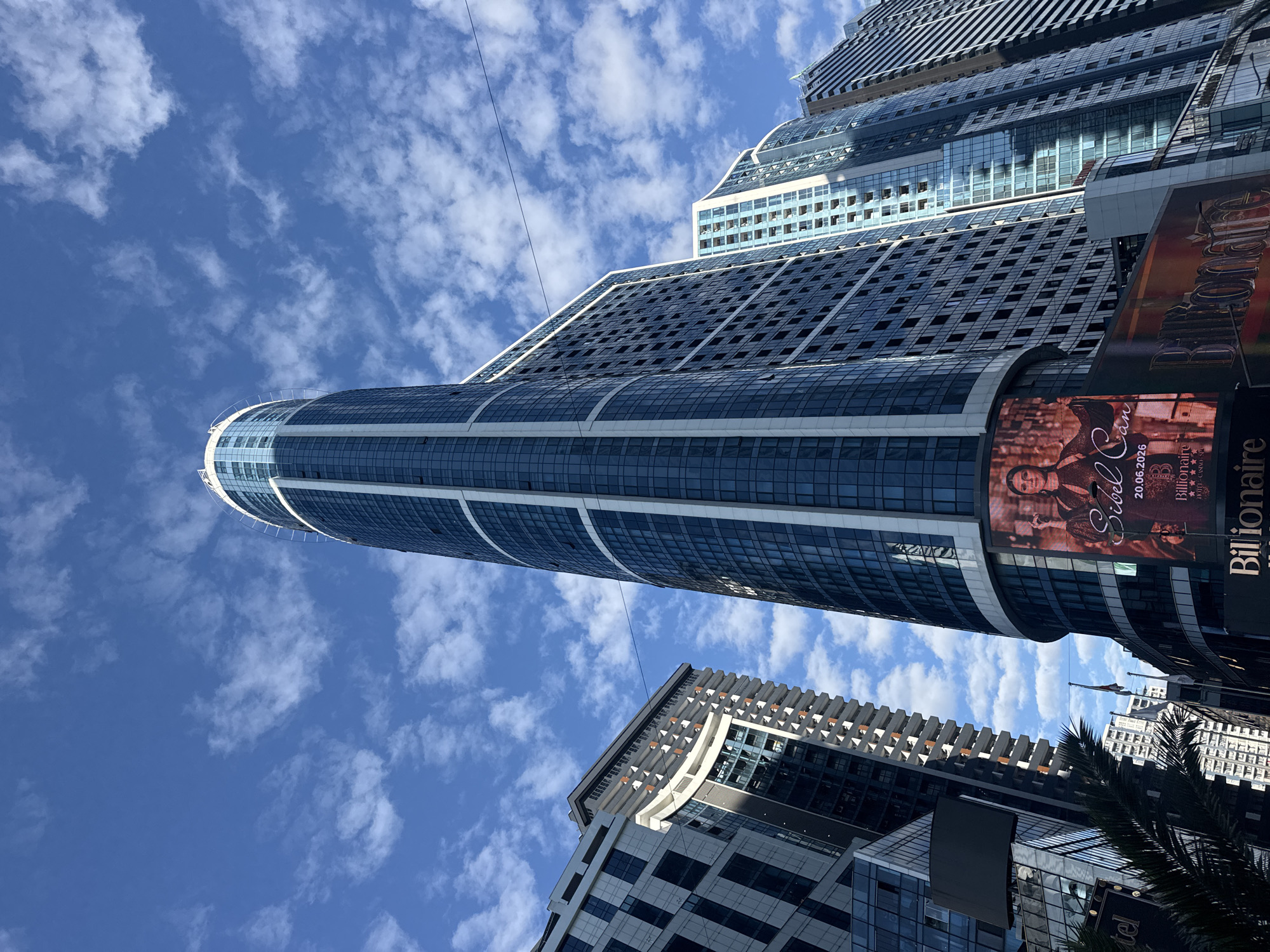 The image, taken with a tilted perspective, captures a vibrant urban scene dominated by modern skyscrapers against a clear blue sky with scattered white clouds. The most prominent building is a tall, slender high-rise with a curved, cylindrical design, featuring a reflective blue glass facade accentuated by white horizontal bands. This building extends from the bottom left towards the top right, appearing to curve slightly out of frame at its apex.
To the right of the central skyscraper, another large, tiered building with a complex structure is visible. It features numerous rectangular windows and darker glass panels, creating a textured appearance. Further in the background, other contemporary high-rise buildings contribute to a dense urban skyline. Thin black lines, possibly power lines or cables, crisscross the sky.
A large digital billboard is mounted on the side of one of the buildings on the right. This billboard displays an advertisement featuring a woman, identified by the text "Sibel Can." Below her name, the date "20.06.2026" is prominently shown. Other visible text includes "Billionaire," and partially visible words like "L'OTEL" and "CASINO," suggesting an entertainment or hospitality complex. A section of a palm tree is also visible in the lower right corner, indicating a warm climate. The scene is brightly lit, suggesting it is daytime under sunny, partly cloudy weather conditions, portraying a bustling city environment.