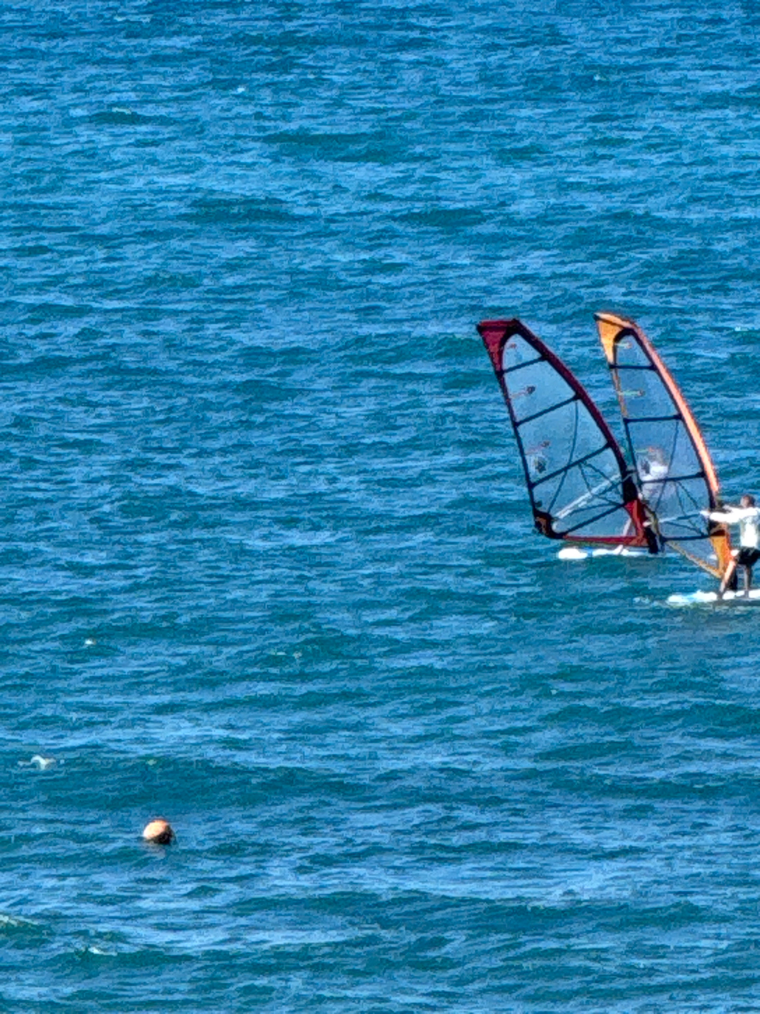 Two windsurfers are on the water. They are both in the process of windsurfing. The windsurfer on the left has a dark red sail. The windsurfer on the right has an orange sail. Both windsurfers are standing on their boards. The sea is blue and choppy. There is a buoy in the distance. The scene is likely located near the coast of Anapa, Russia, as windsurfing is a popular activity in the region.