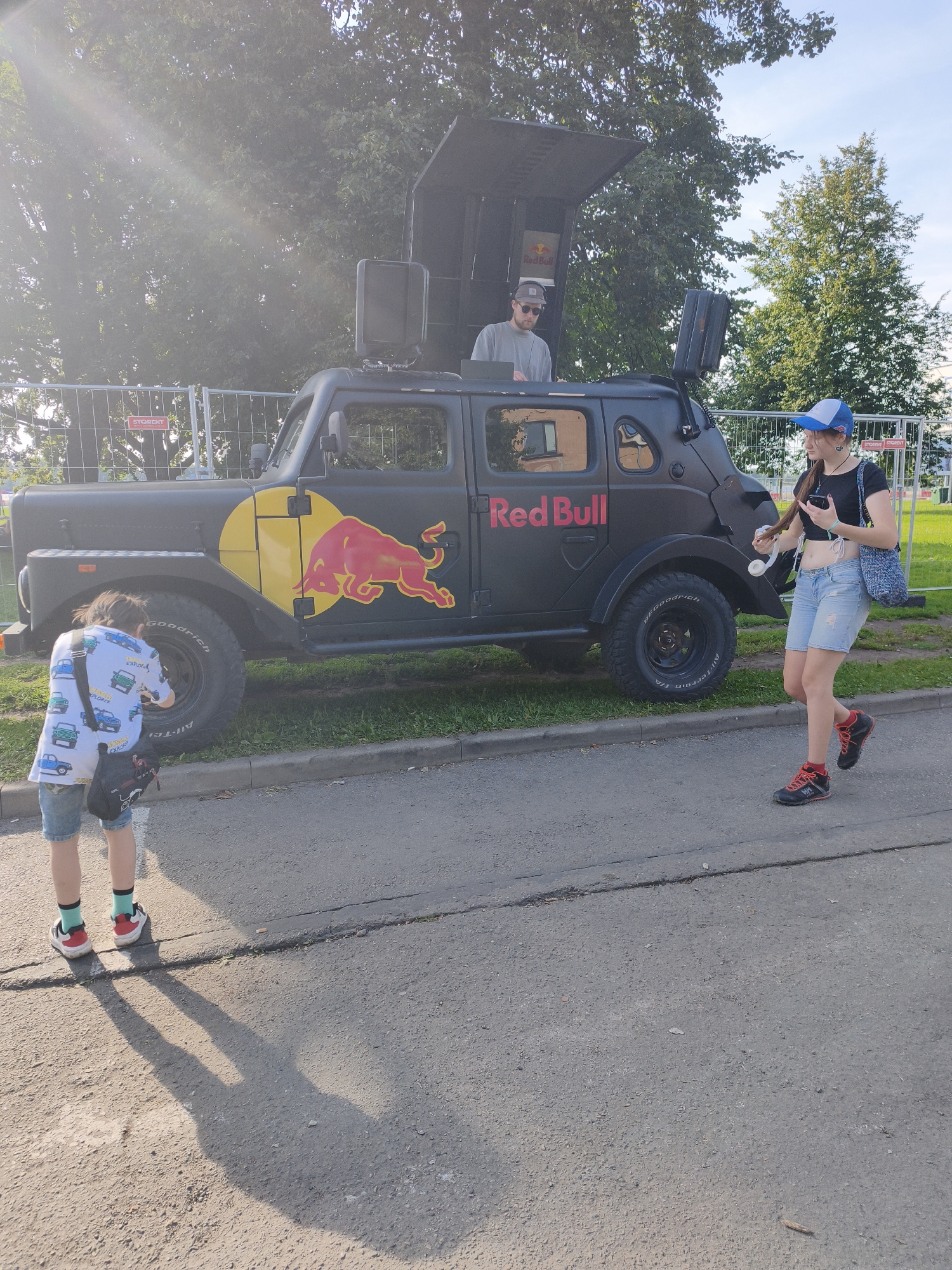 In Riga, Latvia, on a bright, sunny day, a modified, dark-colored vehicle with a raised roof structure is parked on the side of an asphalt road. The vehicle is decorated with a large red bull logo on its side, and the words "Red Bull" are prominently displayed. A person in a grey t-shirt and baseball cap stands in the raised roof section of the vehicle, appearing to be operating equipment. To the right of the vehicle, a young woman wearing a blue and white baseball cap, a black crop top, and denim shorts is walking and looking at her phone. She carries a woven blue bag over her shoulder and is wearing red and black athletic shoes. In the foreground, on the left, a child facing away from the camera is taking a photo of the vehicle with a camera. The child is wearing a short-sleeved shirt with a car print, denim shorts, and white sneakers with red accents. The environment features a grassy area with a fence behind the vehicle and trees in the background. The sun casts distinct shadows on the asphalt.