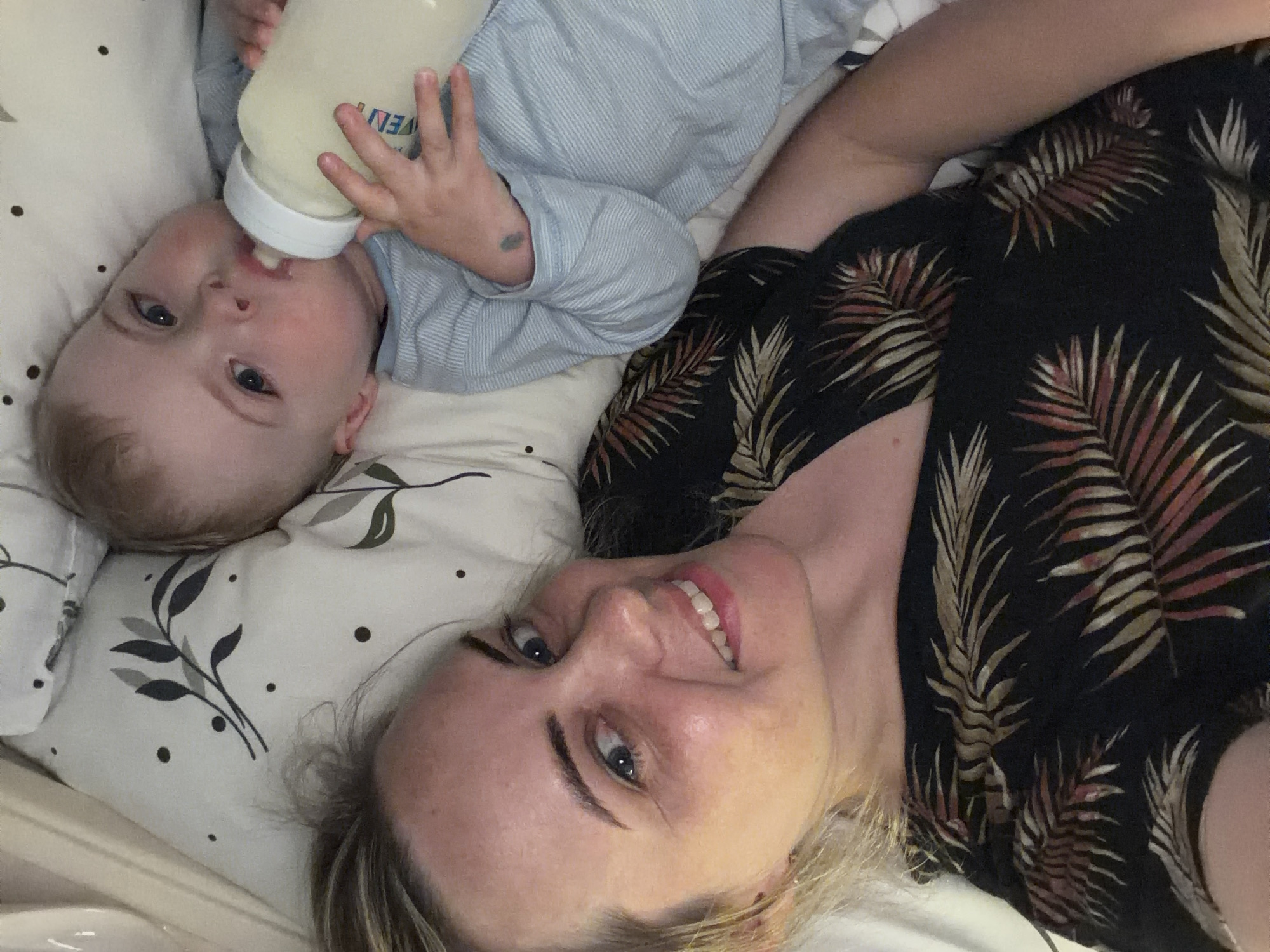 A woman and a baby are lying on a bed with a patterned pillow. The baby, wearing a light blue striped shirt, is drinking from a bottle. The woman, wearing a dark shirt with a leaf pattern, is smiling at the camera.
There is no indication of the location of the photo being Votkinsk, Russia.