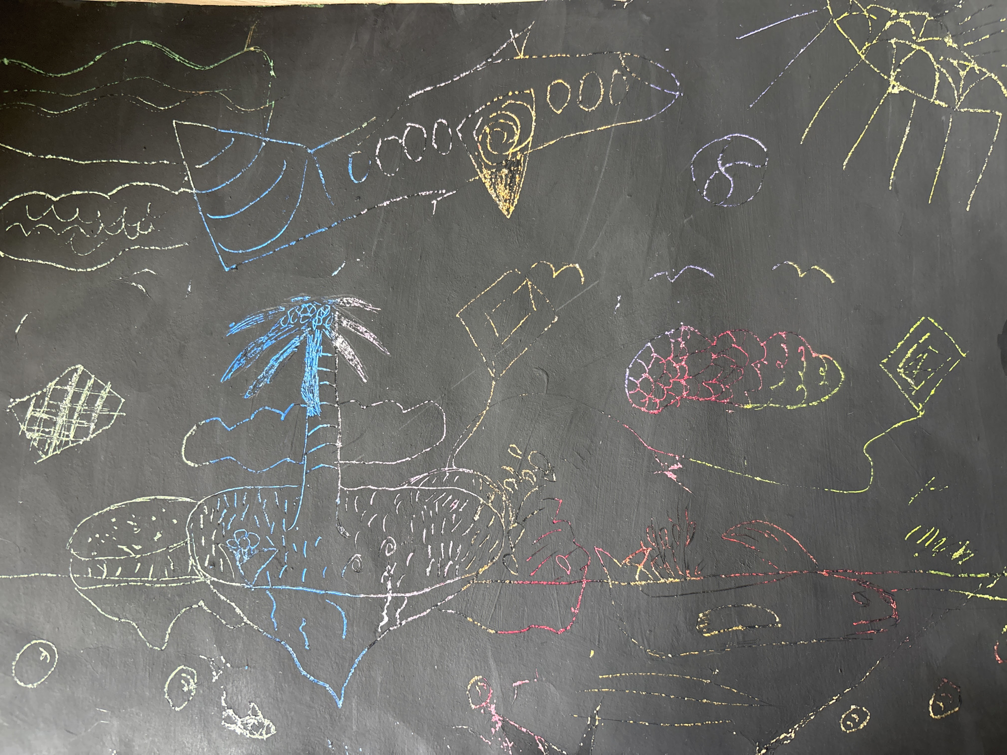 The image is a child's chalk drawing on a black surface. It depicts a tropical scene featuring a palm tree on an island, an airplane in the sky, waves, clouds, and a sun. The drawing includes outlines of shapes such as a building and figures possibly representing people.
There is no indication this is set in the city of Izhevsk, Russia.