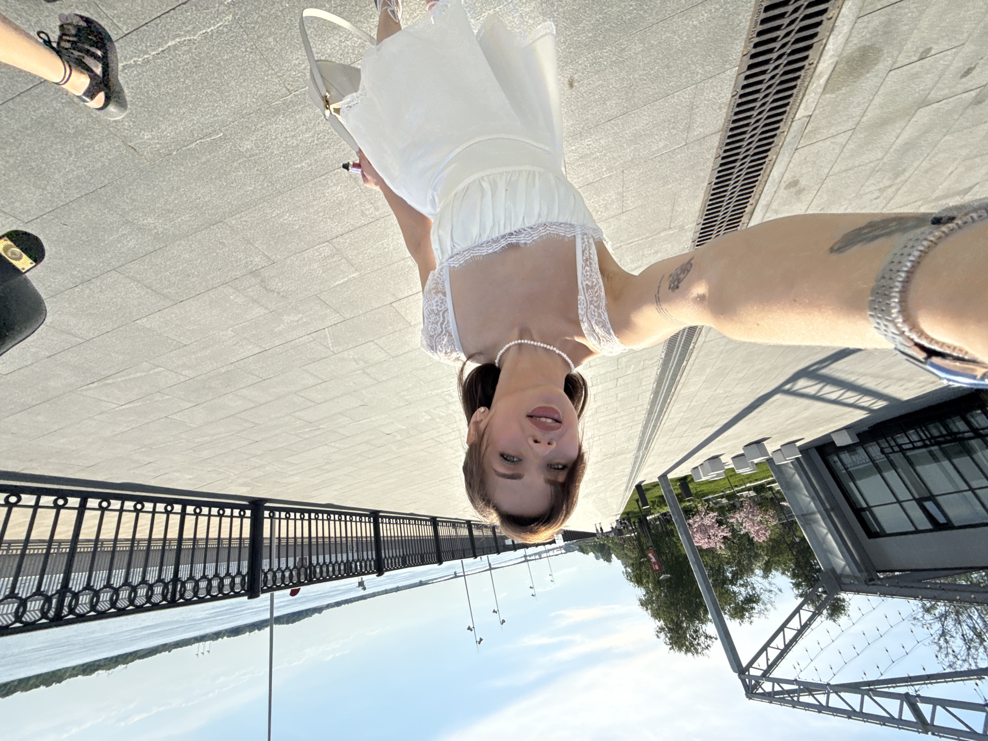 A selfie shows a woman on the Perm embankment of the Kama River. She is fair-skinned, wears a white dress and pearl necklace, and is partially visible in the upper center. Her arm with bracelets is visible on the right. Her feet and a woman's shoes are partially visible in the upper left. The paved embankment is visible as a wide horizontal expanse. A section of the river's iron railing is to the left, with the river and sky beyond. A modern building with a glass facade stands to the right, with trees and green spaces. A metal grating is at the top right of the image.
