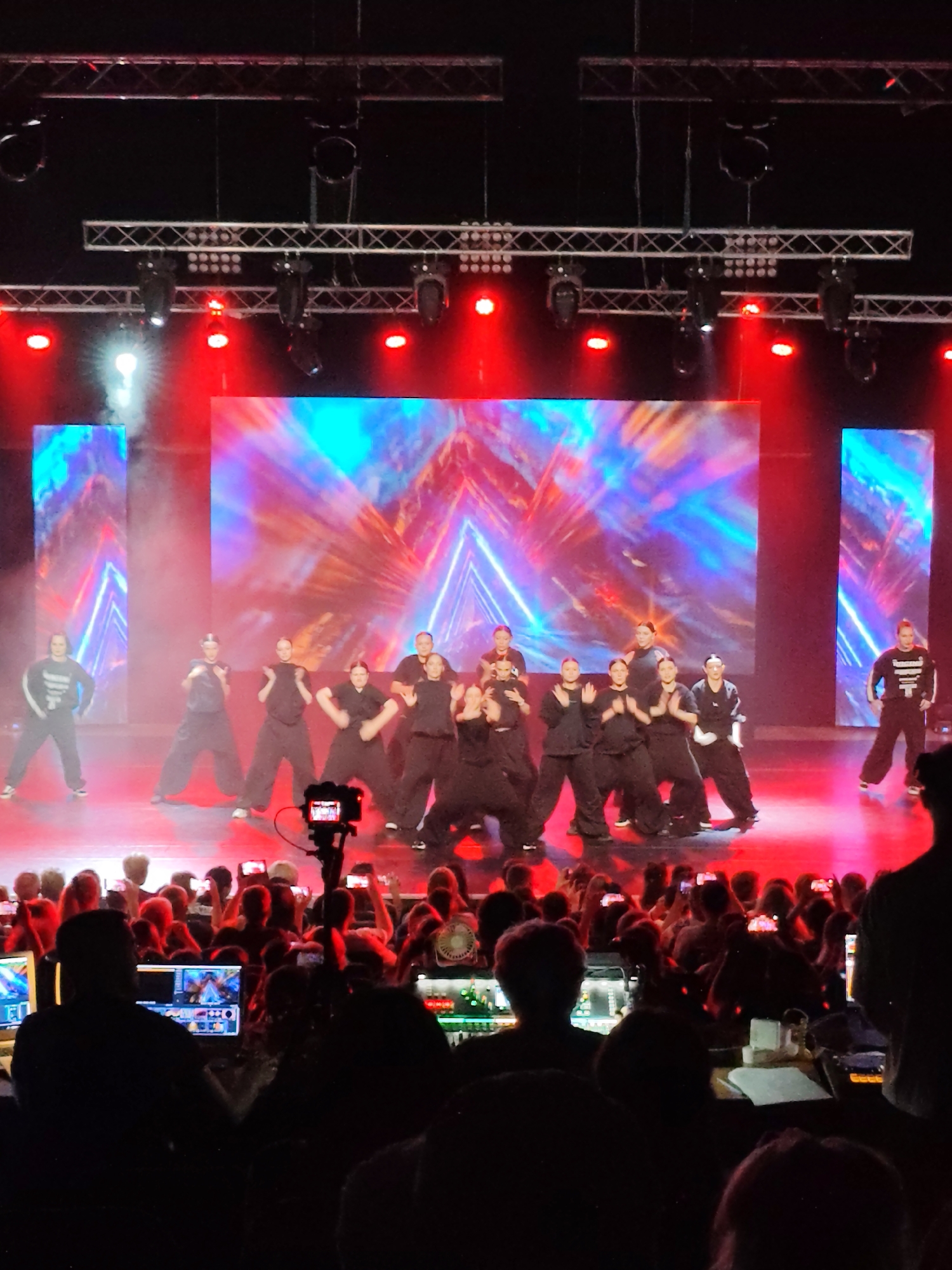 A group of young dancers in black outfits perform on a stage with a vibrant, abstract digital backdrop and red stage lighting. They are positioned in front of two large screens displaying colorful, geometric patterns. In the foreground, the audience is visible, with many individuals holding up smartphones to record the performance. The scene appears to be an indoor event, possibly a dance competition or show, taking place in a venue in Mykolaiv, Ukraine. The overall atmosphere is energetic and celebratory, with the focus on the performers and the engaged audience.