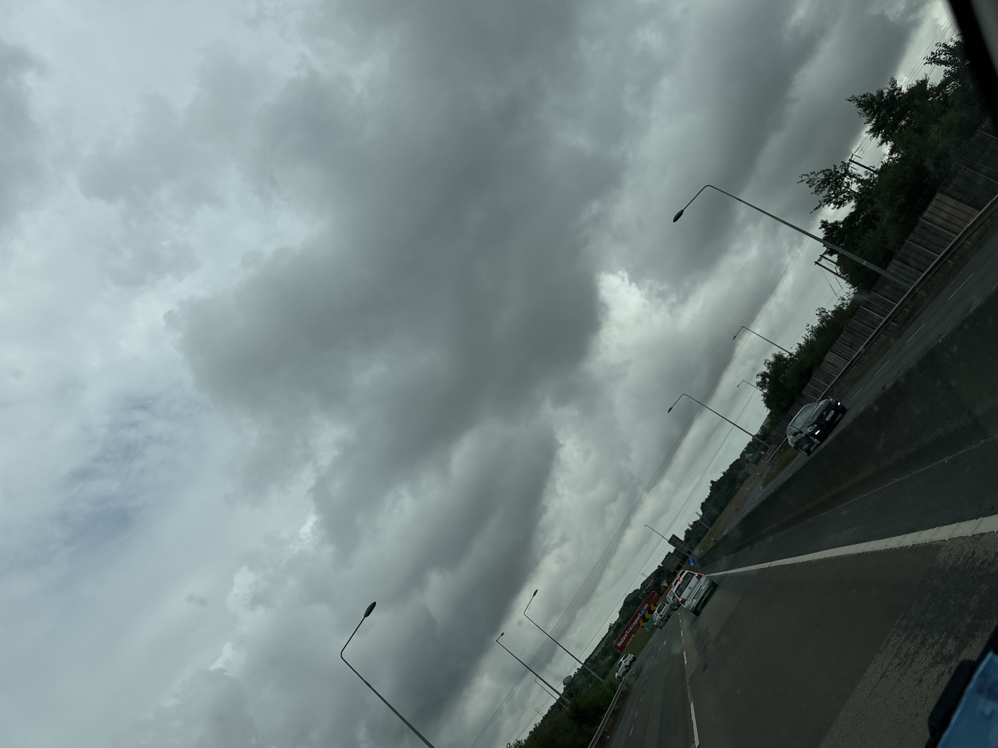 The image captures a view from a vehicle traveling on a highway, likely in or near Slieverue, Ireland, under a heavily overcast sky. The road surface appears wet, suggesting recent or ongoing precipitation. Several cars are visible on the multi-lane highway, indicating moderate traffic. Streetlights line the road at regular intervals, their lamps illuminated against the dim conditions. Vegetation lines the side of the highway, with trees and shrubs forming a green barrier. A large billboard with red text, possibly advertising "NolanTransport.com", is visible in the distance. The overall atmosphere is moody and somber due to the dark, cloudy sky, which dominates the upper portion of the frame. The perspective is angled, giving a dynamic feel to the scene of transit.