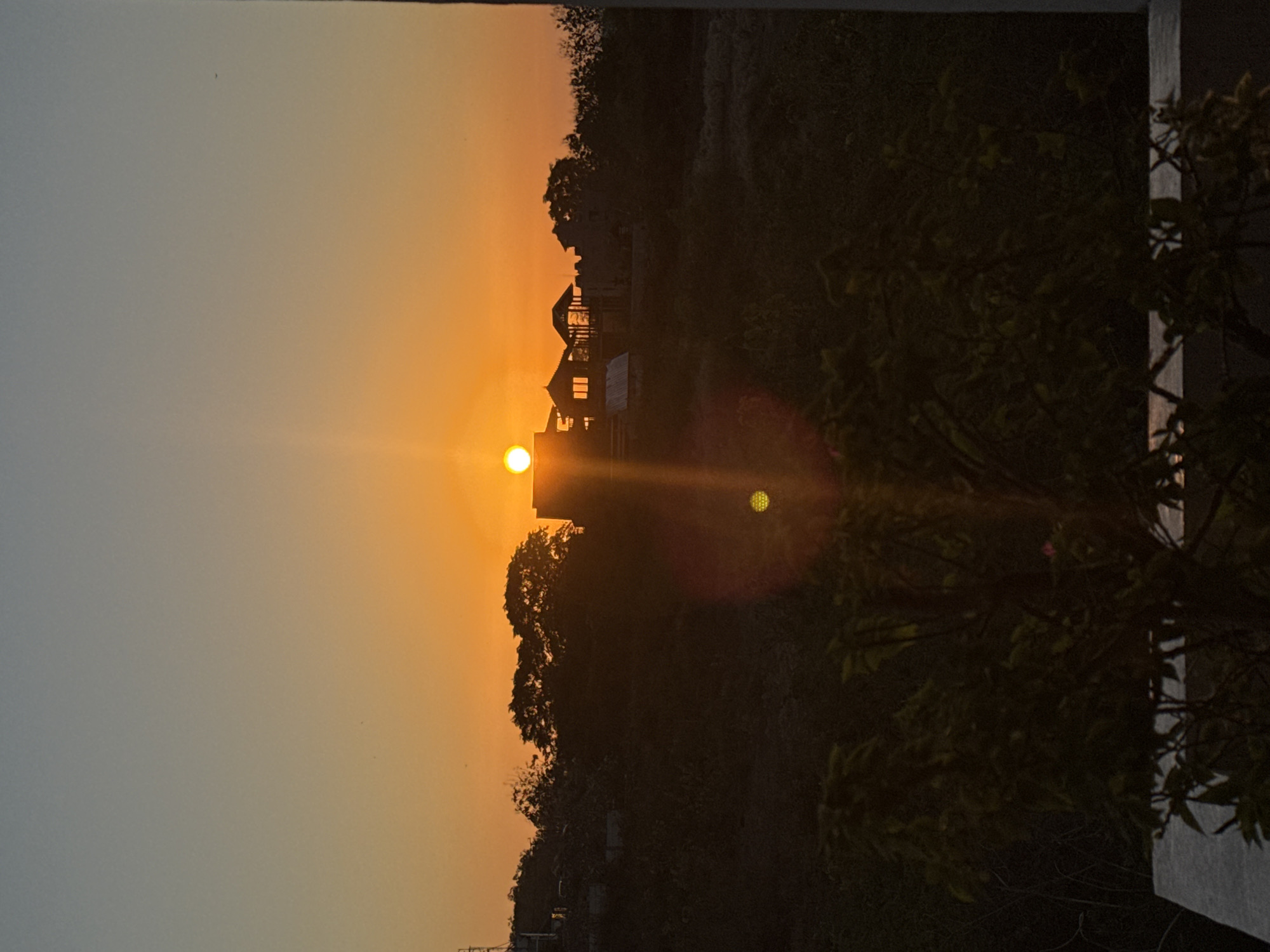 The image shows a sunset over Kuta Selatan, Indonesia. The sun is setting behind a silhouetted line of trees and buildings on a hill. The sky is a gradient of orange and yellow, with a few birds flying in the distance. The foreground is dark and out of focus, with some leaves and a concrete structure visible. The overall mood is serene and peaceful.
