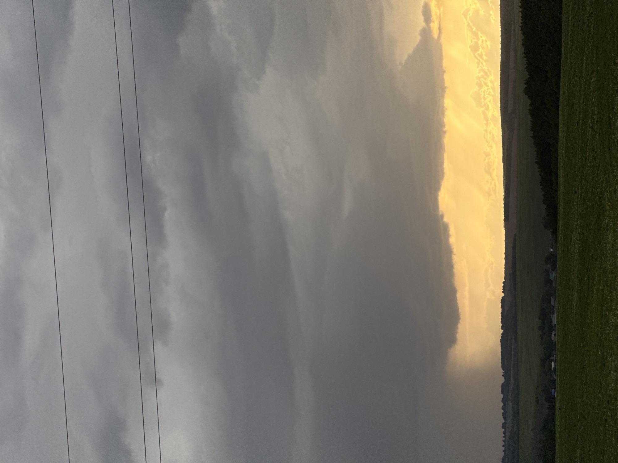 The image shows a landscape under a dramatic sky. In the foreground, power lines are visible against a backdrop of heavy, grey storm clouds. A contrasting patch of bright, yellow light illuminates a portion of the sky and possibly a downpour of rain, positioned over what appears to be a field. The right side of the image includes a dark treeline and a green field. Small structures, possibly buildings, are discernible near the treeline. There is no confirmed information about people being present in the image. The location in the city of Крынды, Russia, is also unconfirmed.