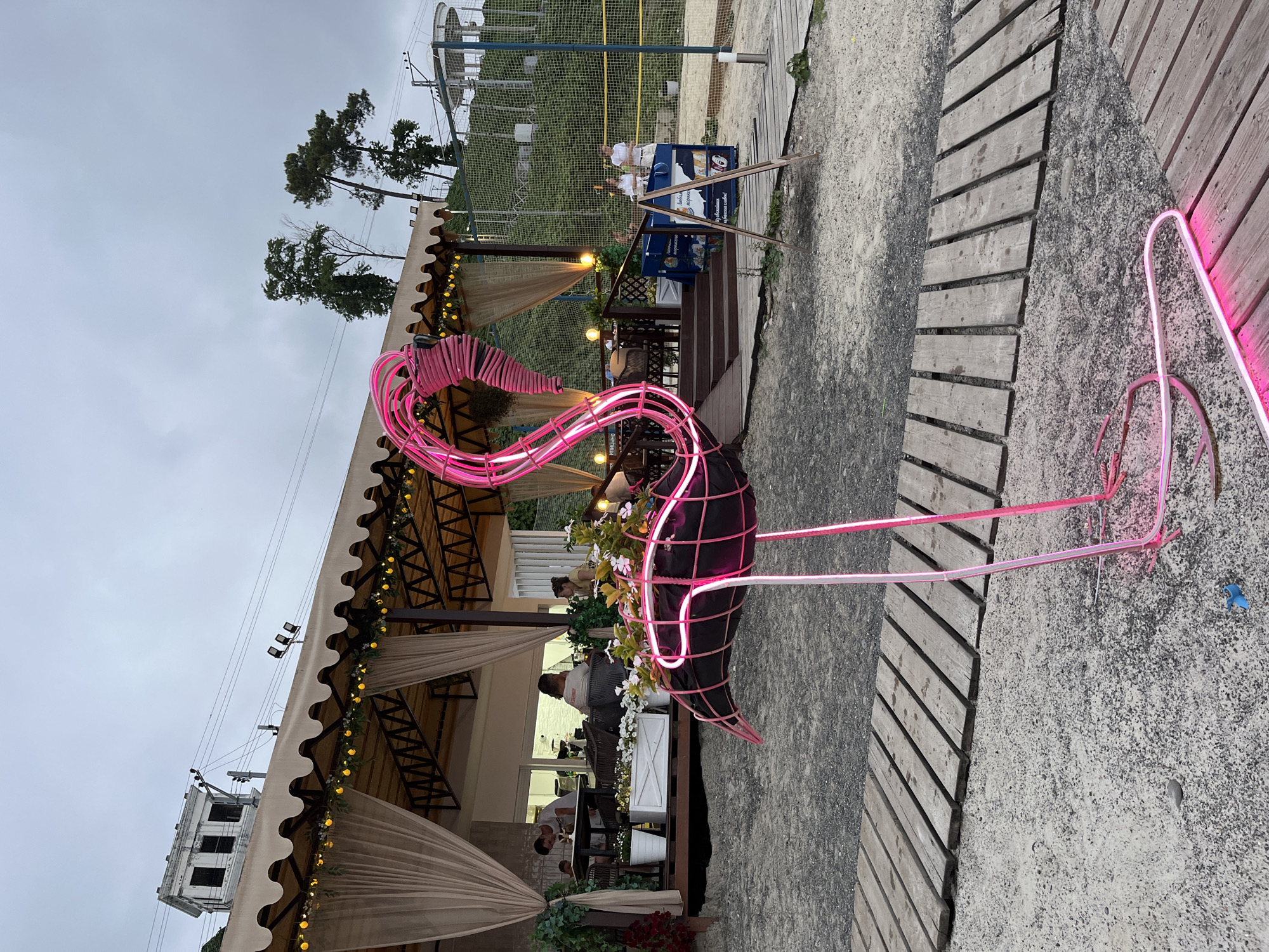 This image depicts an outdoor cafe or restaurant area, possibly on a beach or near a body of water, located in Sochi, Russia. In the foreground, a vibrant neon flamingo sculpture, primarily pink, stands on thin legs. The flamingo is dark-colored within its neon outline and appears to be holding a planter with white flowers. Wooden planks form a walkway in front of the sculpture.
Behind the flamingo, a covered outdoor seating area is visible. The structure has a beige awning with scalloped edges, supported by dark metal beams. Yellow string lights are draped along the awning. Inside the covered area, tables and chairs are set up, and several people are seated, dining or conversing. Two servers, a woman and a man, are visible near the entrance, one of whom is placing an order.
Further in the background, a green, grassy hill rises, topped with trees and some buildings. A white building with dark windows is prominent on the upper left side. A sports court, possibly a volleyball court with a net, is also present in the background, with a few more people milling around. The sky is overcast, suggesting a cloudy day. The overall atmosphere appears to be relaxed and casual, typical of a seaside establishment.
