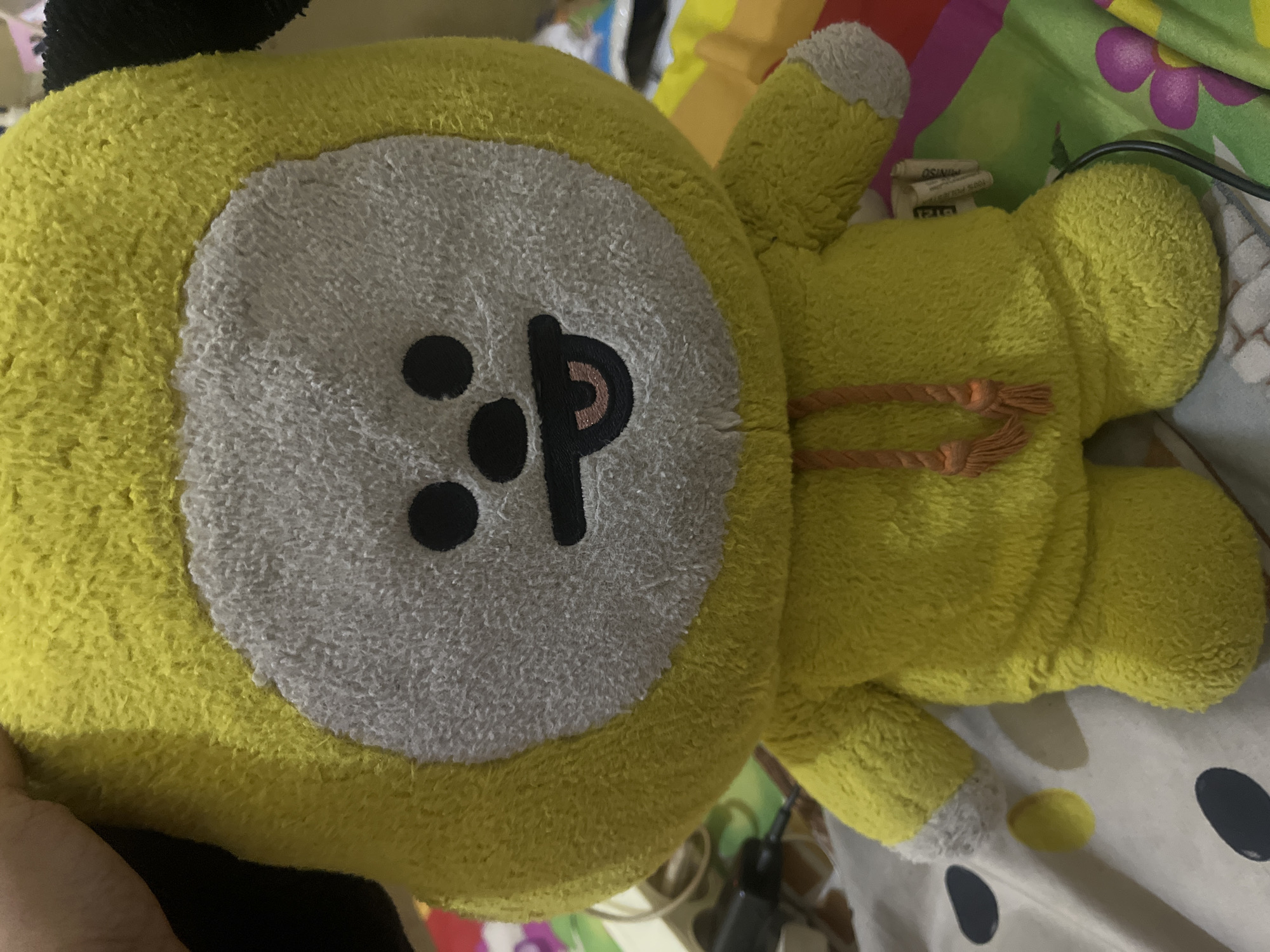 The image shows a yellow Chimmy plush toy lying on a patterned surface. The Chimmy plush has a white face with black eyes and a black mouth in the shape of the letter 'p'. The plush toy also has two orange straps attached to its back. The background includes a colorful patterned surface with purple flowers and a black power adapter. The image was likely taken indoors. There is no information in the image to confirm the location in Central Jakarta, Indonesia.