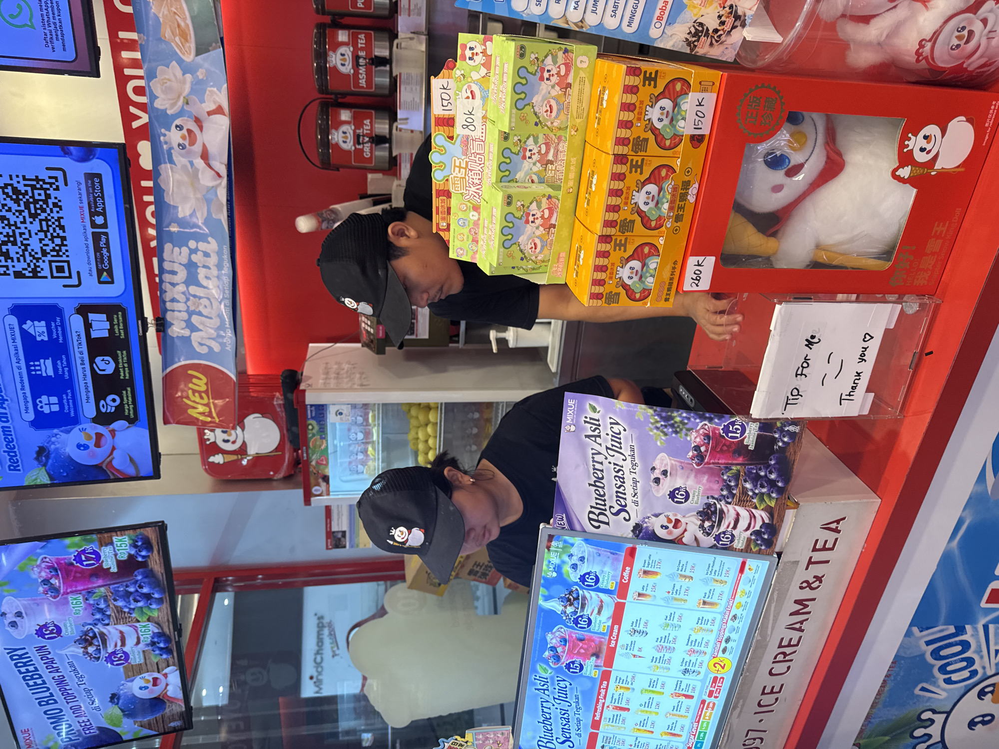 This image depicts the interior of a Mixue ice cream and tea shop in Kuta, Indonesia, likely a franchise location given the branding. The scene is brightly lit, with red and white being dominant colors. In the foreground, a digital menu board displays various ice cream and tea options with prices in Indonesian Rupiah (Rp). Two employees are visible behind the counter. One is wearing a black Mixue branded cap and a black t-shirt, looking down at the menu board. The other employee, partially visible, is also wearing a black cap.
On the counter, stacked boxes of merchandise, including what appear to be collectible snowmen figures and snack boxes, are displayed. Some boxes have prices labeled on them, such as "150k" and "80k". A large red box with a clear window reveals a stuffed snowman toy. To the right, a white sign with "Tip For Me" and a hand-drawn smiley face and heart suggests a tip jar.
In the background, shelves with various drinks and packaged goods are visible. A "NEW" sign is prominently displayed on a red backdrop. The overall impression is a busy retail environment focused on selling ice cream, tea, and related merchandise. The signage and menu prices indicate the location is in Indonesia.