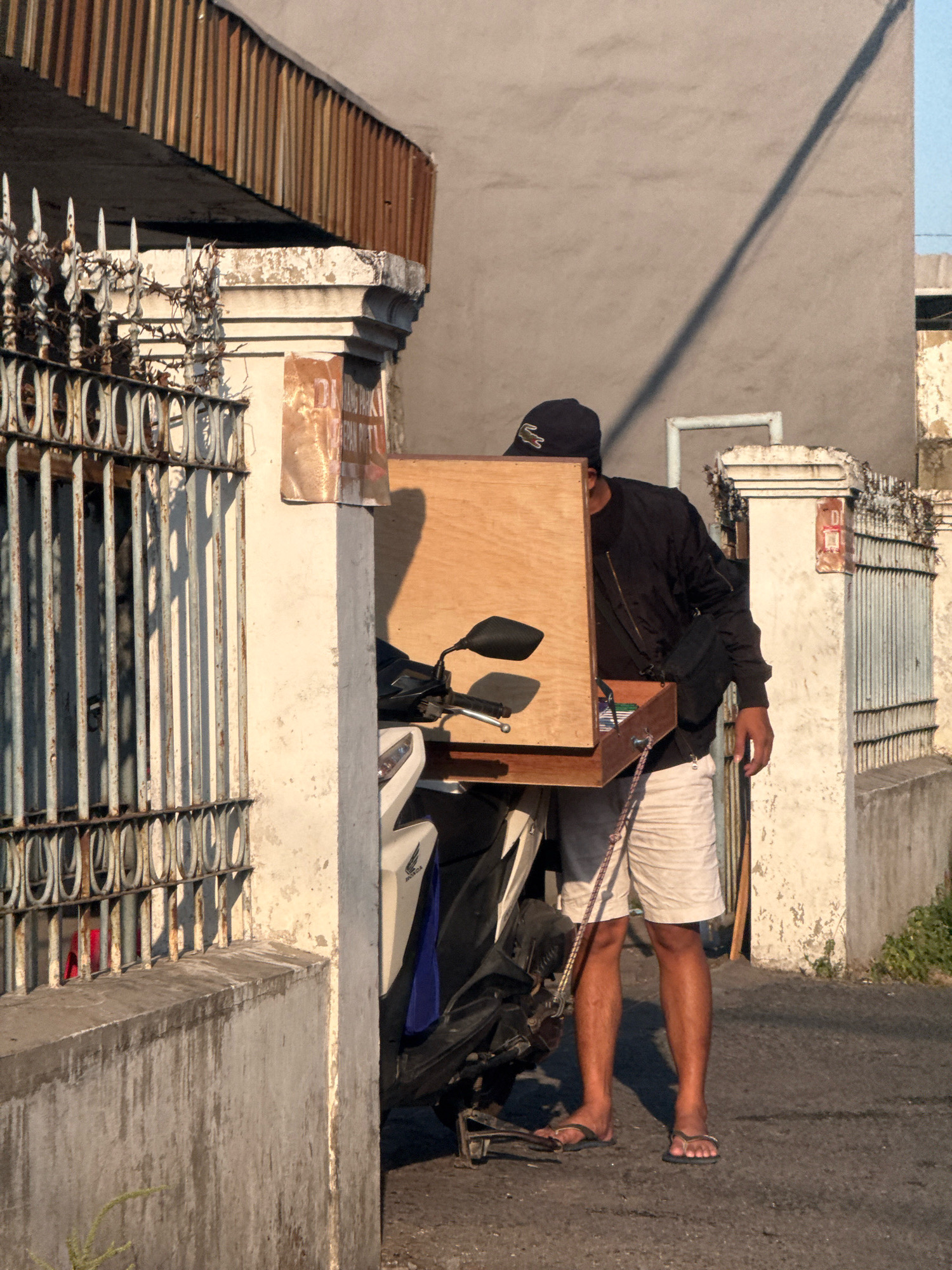 A person stands on a paved street next to a parked motorcycle. The person, wearing a black baseball cap, black jacket, and light-colored shorts, is partially obscured by a wooden box attached to the motorcycle. The box appears to be a makeshift carrier. The motorcycle is white and black, with a blue fabric covering part of it. The background includes a concrete wall with a faded advertisement, a metal fence with decorative elements and some rust, and a building with a slatted overhang. The scene is illuminated by sunlight, casting shadows. The overall environment suggests an urban or semi-urban setting in Surabaya, Indonesia.