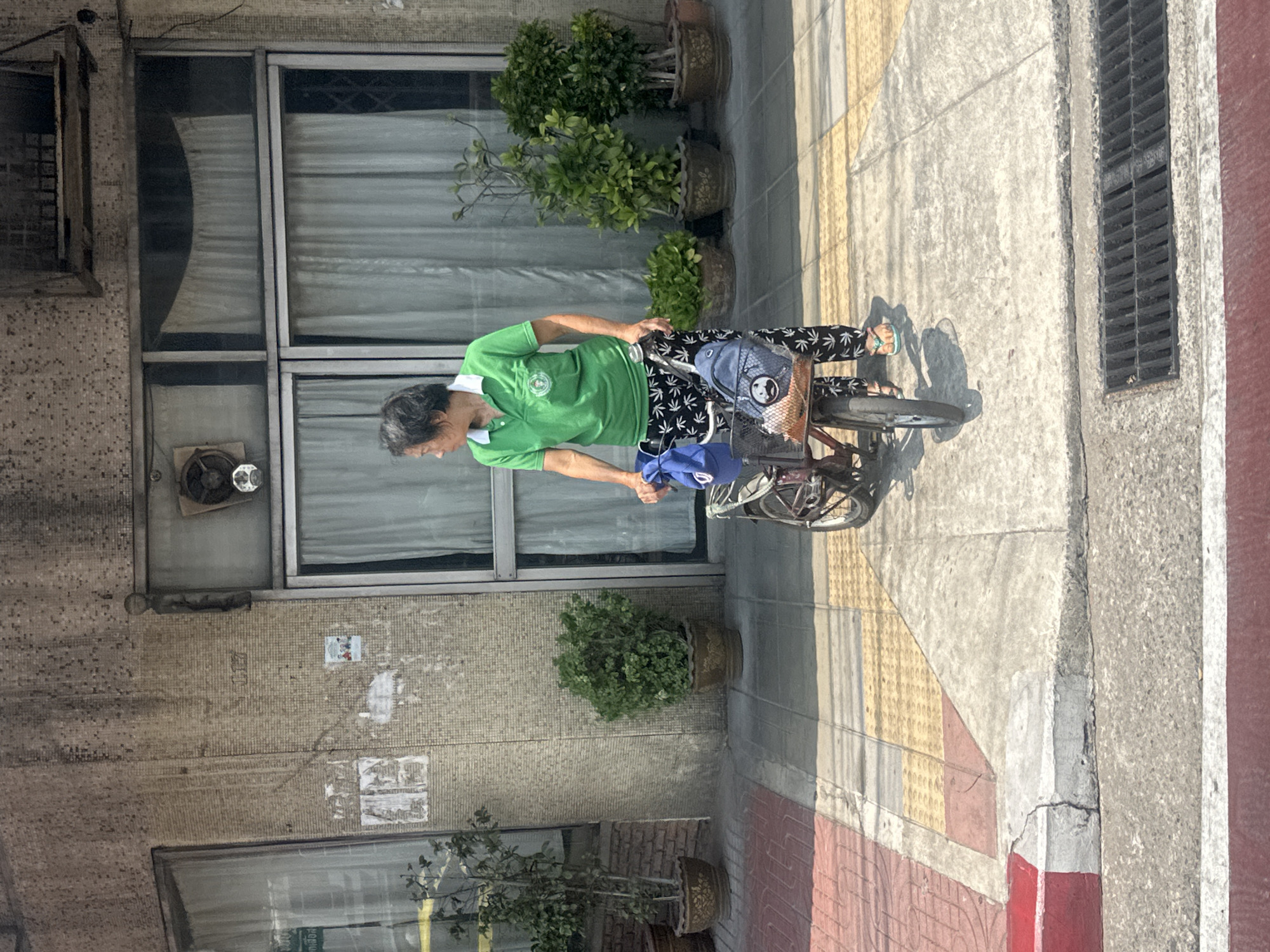 An older Thai woman in a green shirt and marijuana-leaf patterned pants is riding a bicycle on a sidewalk in Bangkok, Thailand. She has a blue bag and a blue hat in the bicycle's basket. The sidewalk is partially covered with paving stones in a pattern of yellow, red, and grey colors. Behind her, a building with aging architecture has a series of windows covered with curtains, and potted plants sit on the sidewalk in front of the building.
