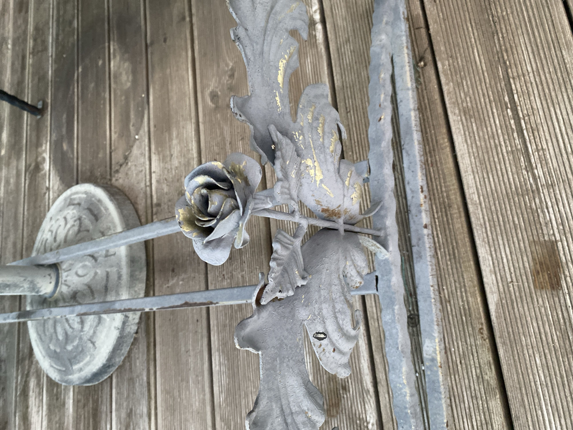 The image shows a weathered metal garden ornament with a rose and leaf motif, placed on a wooden deck. The ornament is painted a muted gray with hints of gold highlighting its details. The wooden deck appears to be made of planks laid horizontally, showing signs of age and exposure to the elements. In the background, a circular, textured base of what appears to be a metal stand or table is visible, also on the wooden deck. The scene suggests an outdoor setting, possibly a garden or patio area, in Bol'shoe Zaborod'e, Russia. There are no people or animals present in the image.