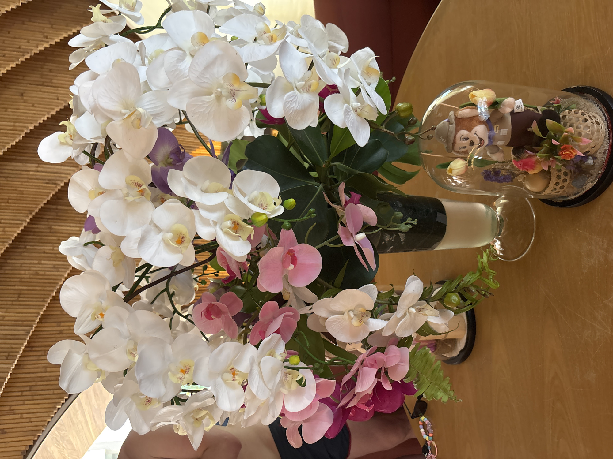 A high-angle shot reveals a floral arrangement primarily consisting of white and pink orchids, displayed in a black vase. To the right, under a glass dome, is a stuffed animal surrounded by artificial flowers and foliage. The arrangement sits on a table in Baan Bo Phut, Thailand. A portion of a light brown, horizontally-striped background is visible. Human skin is visible in the lower center of the frame.