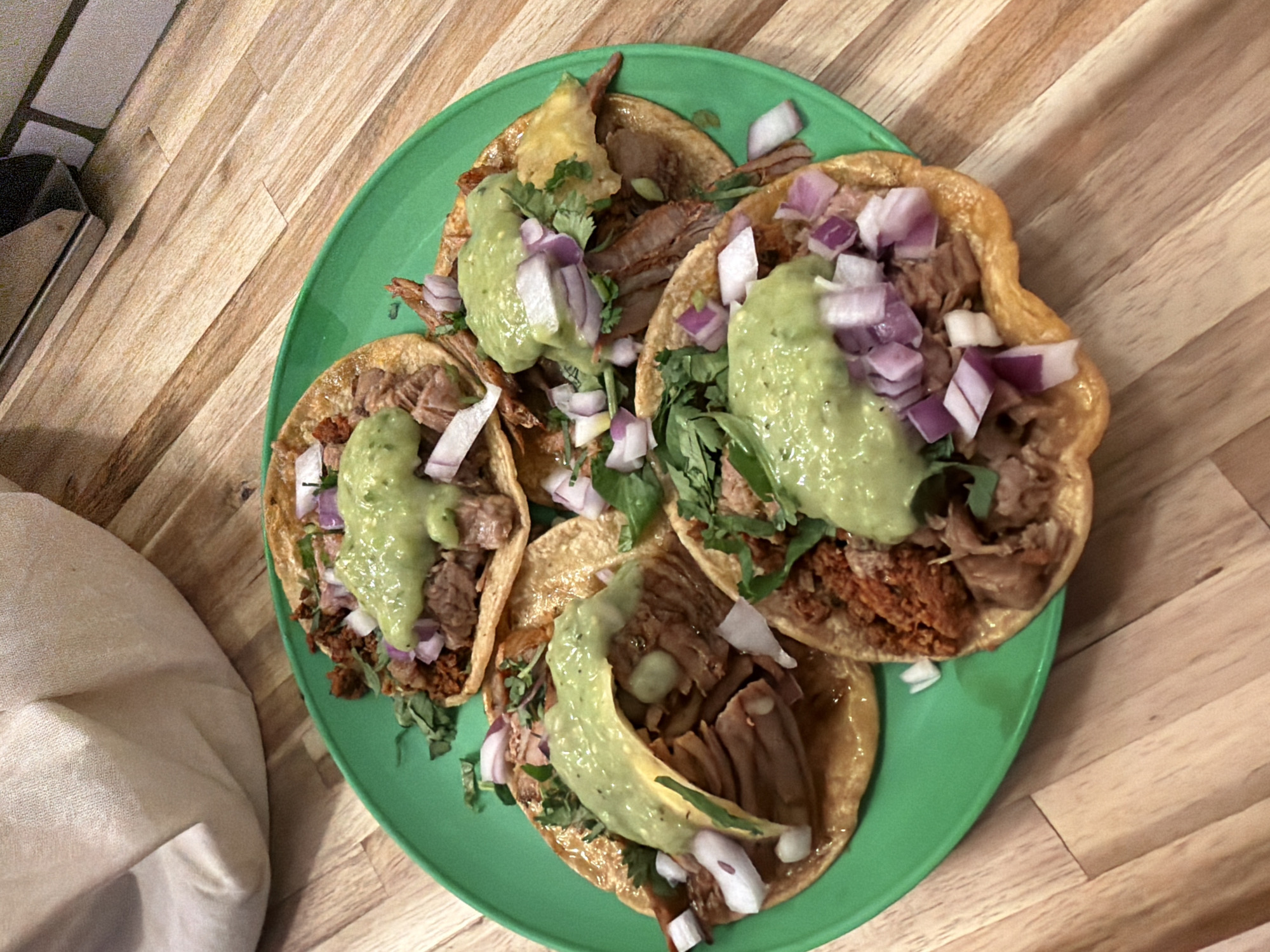The image shows a green plate with four tacos. The tacos are filled with meat, green sauce, red onion, and cilantro. The plate is on a wooden surface. It is not possible to determine the location from the image. The city of New York, United States of America cannot be determined from the image.