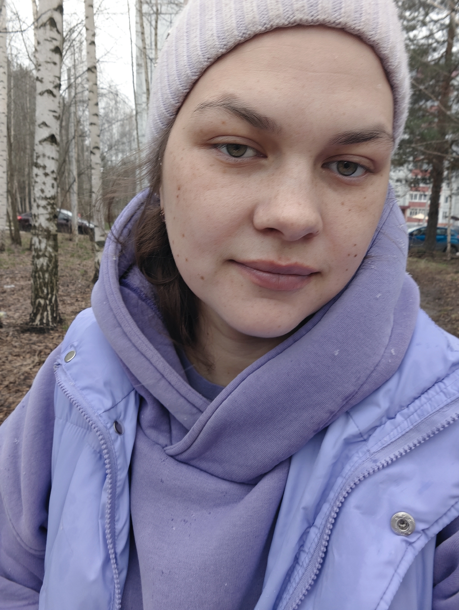 A close-up photograph shows a fair-skinned person with brown hair and green eyes wearing a light purple knitted hat, a purple hooded sweatshirt, and a light purple jacket. The person is looking directly at the camera with a neutral expression. Their face has some freckles. The background includes birch trees with bare branches, some leafless ground cover, and distant buildings and parked cars. The location appears to be a park or wooded area within the city of Chaikovsky, Russia.