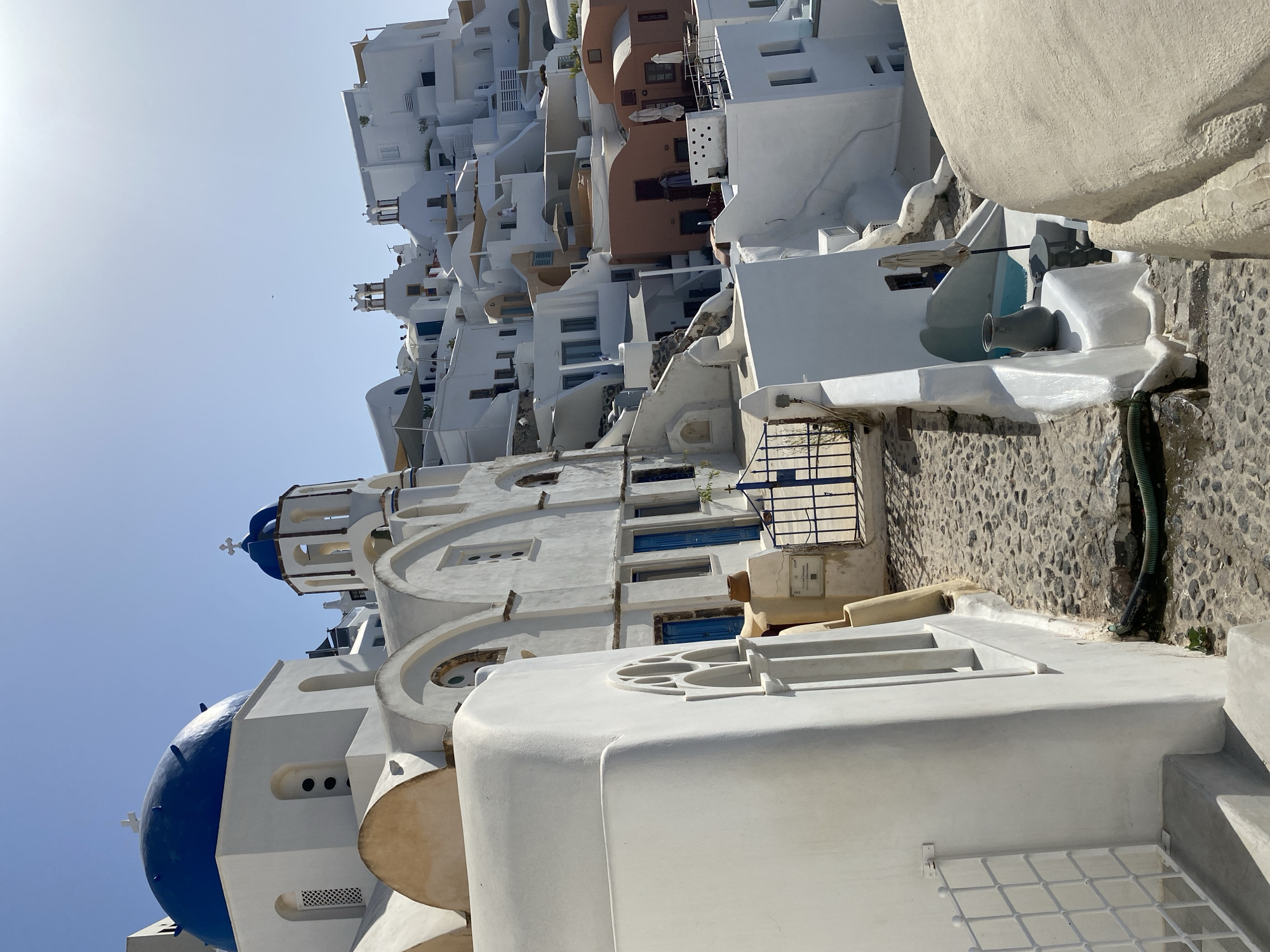 The image depicts a view of the iconic white-washed buildings and blue-domed churches characteristic of Oia, a municipal unit in Santorini, Greece. The scene is dominated by the stark white architecture, contrasted with the vibrant blue of a church dome in the foreground and another in the mid-ground. The buildings are tightly packed together, cascading down a hillside under a clear, bright blue sky. Cobblestone pathways wind through the settlement, with stone steps and narrow passageways visible. Blue-painted doors and window frames add pops of color to the predominantly white structures. Some buildings feature arched windows and decorative elements. A metal gate, also painted blue, is visible on a pathway. In the mid-ground, a small swimming pool is partially visible, with a grey vase and a small table beside it. The architecture suggests a sunny, Mediterranean climate. There are no people clearly visible in the image.