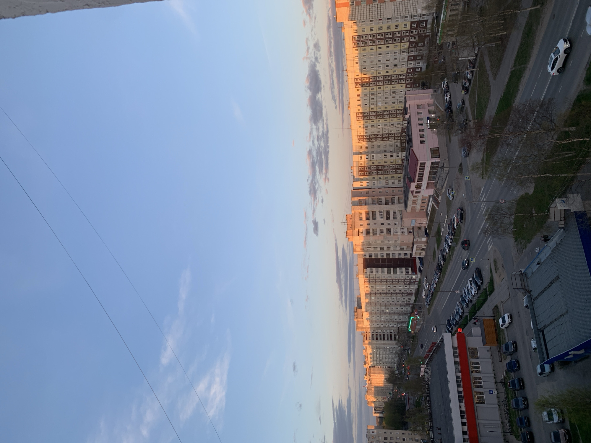 The image presents a high-angle view of a city street in Severodvinsk, Russia, likely captured from a high-rise building. The sky is a gradient of blue, transitioning into lighter hues, indicative of either dawn or dusk, with thin scattered clouds tinged with a faint golden hue. Rows of multi-story residential buildings, typical of Soviet-era architecture, line the street. The buildings are primarily beige or gray, with repetitive window patterns. Some buildings have architectural details or color accents. A street runs parallel to the buildings, showing cars parked along the side and moving traffic. There are sidewalks and pedestrian walkways bordered by rows of trees. The general impression is that the scene is a modern residential area.