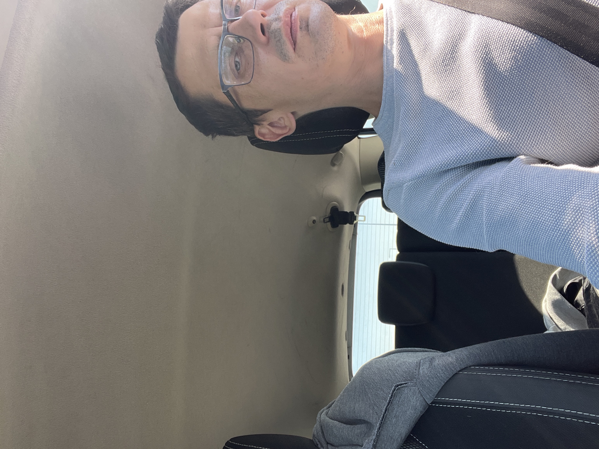 A man wearing glasses and a light blue sweater is sitting in the driver's seat of a car, looking forward. Sunlight streams in from the right side of the image, casting shadows. The car interior is visible, including the headrests of the seats behind him and the car's ceiling. The man appears to be in Zabrze, Poland.