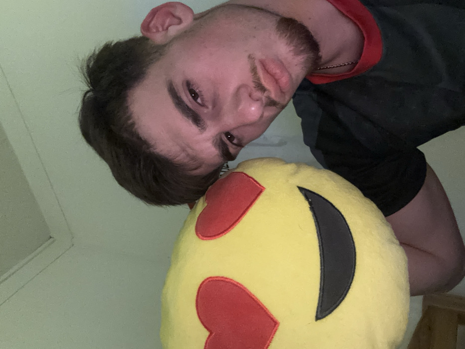 A light-skinned man with dark hair and a short beard is angled in the frame with a pouting expression. He wears a black shirt with a red collar trim and a silver necklace. A yellow emoji pillow with red heart eyes and a black smile sits below him. A white wall is visible in the background, with what appears to be a window frame on the left. The image appears to be taken at an upward angle.