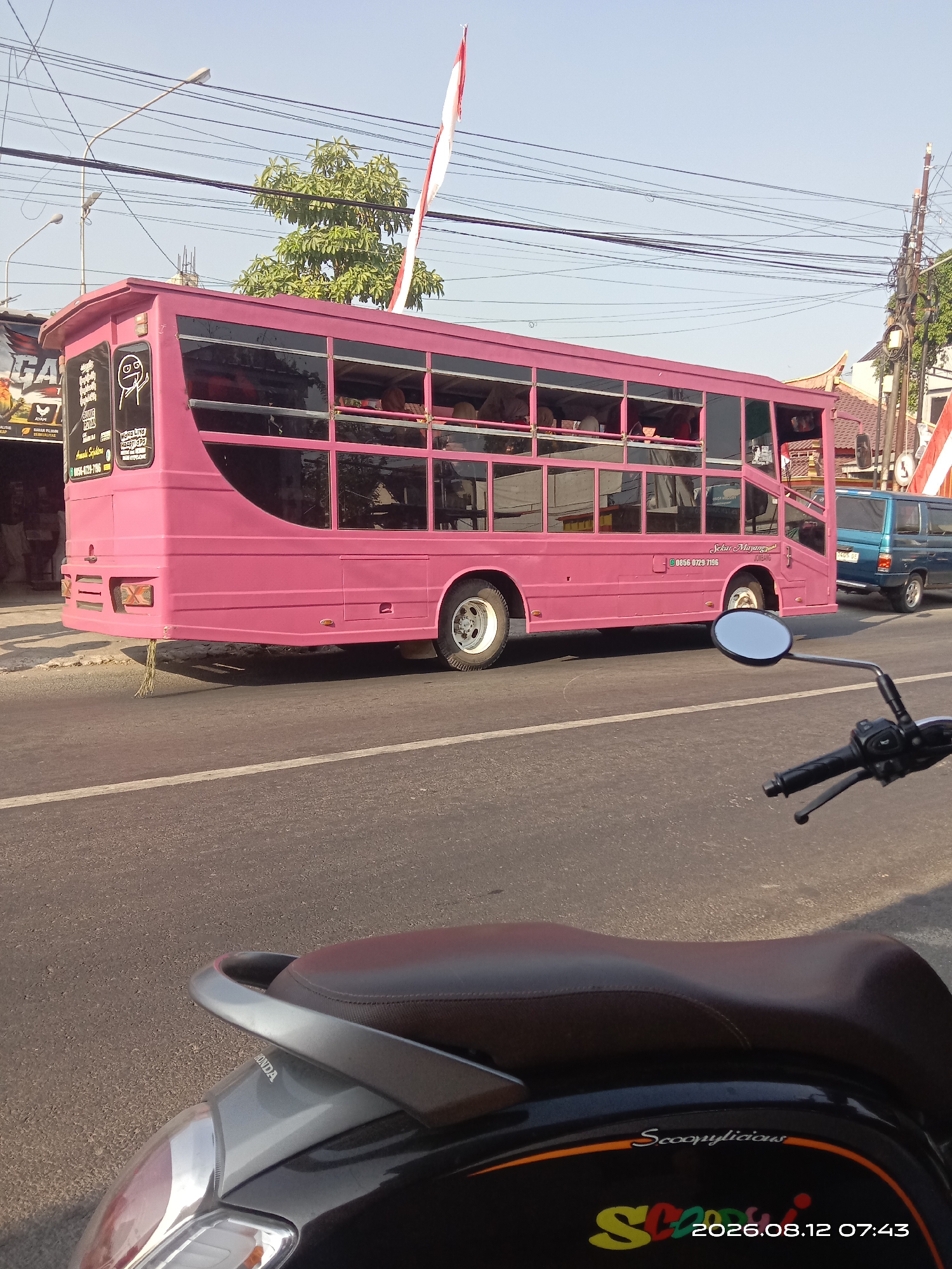 In Gudo, Indonesia, a bright pink double-decker bus travels along a paved road under a clear sky. The bus features large tinted windows, revealing passengers inside, and is adorned with decals and writing. One decal shows a cartoon character with a distressed expression. Text on the bus includes "SEKAR MAYANG" and a phone number. A bright red and white Indonesian flag is visible on a pole atop the bus. To the right, a blue van follows closely behind. In the foreground, the handlebars and brown seat of a scooter are visible, suggesting the photograph was taken from a moving vehicle. The road has a white dividing line, and power lines crisscross overhead. Trees and buildings line the side of the road.