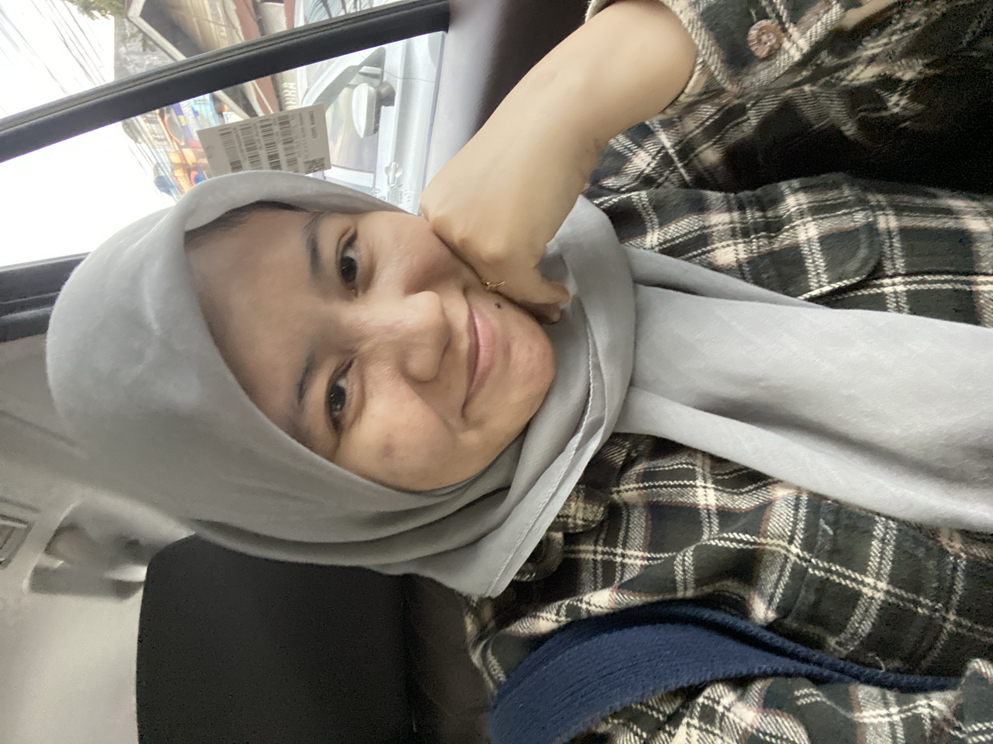 A woman wearing a grey hijab and a plaid shirt is taking a selfie in a car. She is smiling and resting her chin on her hand. A blue bag is visible on her left shoulder. Outside the car window, buildings and a street can be seen. The image appears to have been taken during the day.