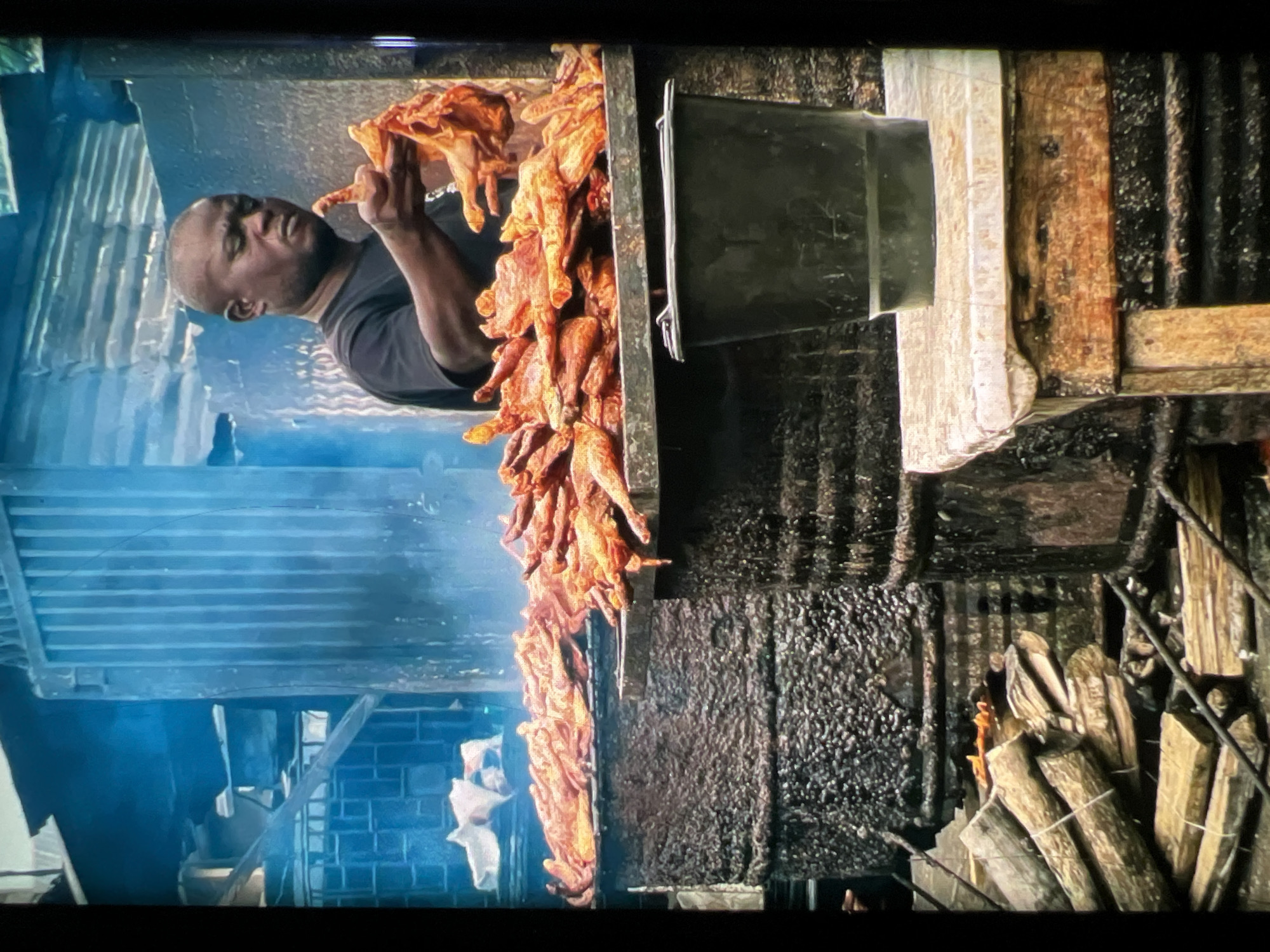 The image depicts an outdoor food stall in Lagos, Nigeria, likely engaged in the preparation of grilled meat. A man, wearing a black t-shirt, is centrally positioned, holding several pieces of partially cooked, reddish-brown meat, possibly chicken, in his hands. He appears to be inspecting or arranging them. In the background, a large quantity of similar grilled meat is piled on a metal grate atop a large, dark, cylindrical structure, suggesting it is a cooking vessel or smoker. Smoke is visible rising from this area, indicating active cooking. In front of the stall, a stack of firewood is neatly arranged, ready for use in the cooking process. A dark-colored bucket sits on a wooden stool to the right of the frame. The overall environment appears somewhat industrial, with corrugated metal sheeting and other utilitarian structures visible. The lighting is dim, with a blueish tint, possibly from artificial lighting or atmospheric conditions.