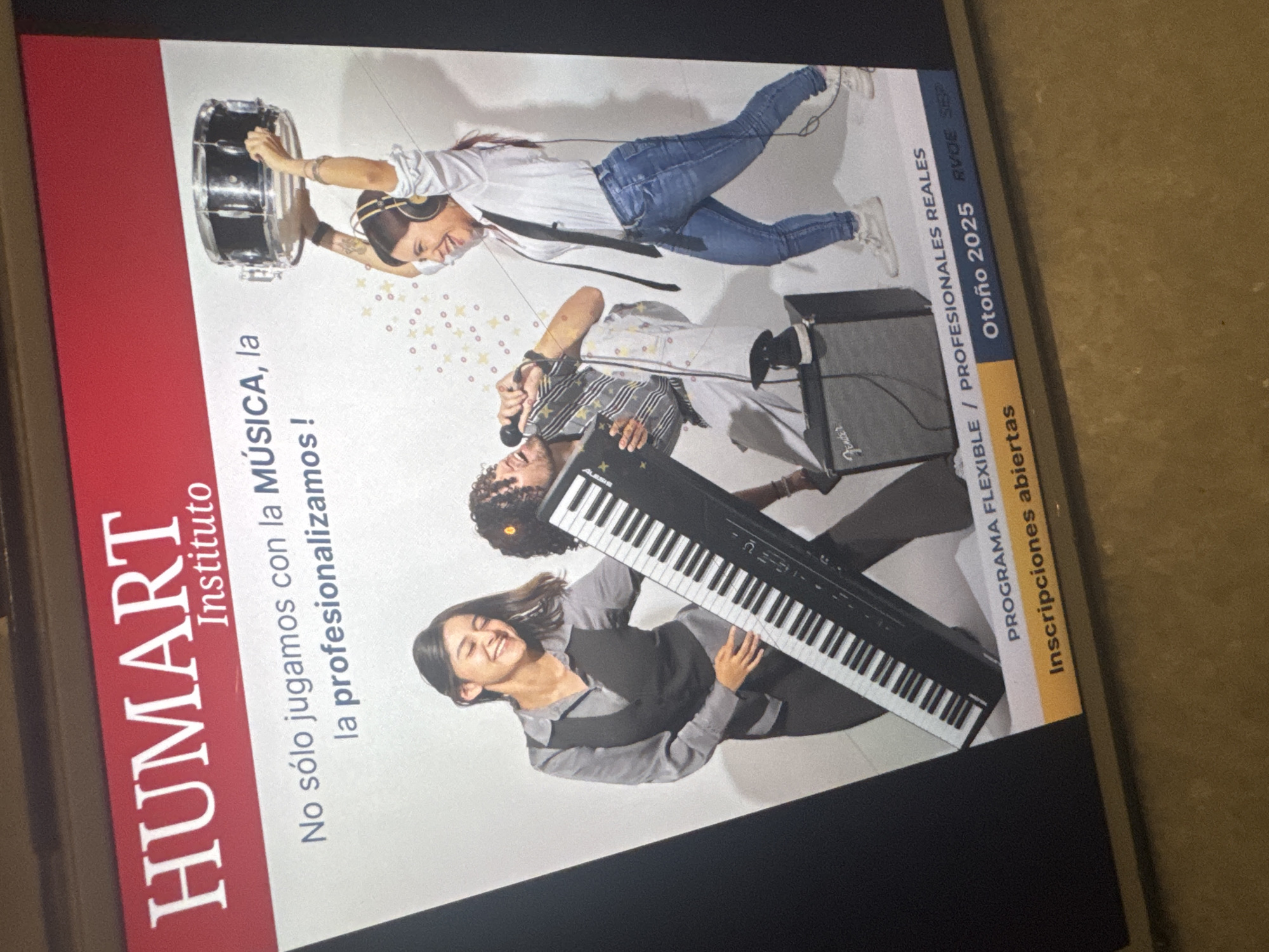 The image is a photograph of a promotional poster for "HUMART Instituto," a music school, likely in Puebla, Mexico. The poster features the school's name in large, red letters on the left side, followed by the tagline "No sólo jugamos con la MÚSICA, la profesionalizamos!" The right side of the poster shows a group of three young people playing musical instruments. One person is playing drums, another is singing into a microphone while playing a keyboard, and the third person is playing a piano. The poster also contains text indicating "Otoño 2025" and the phrases "PROGRAMA FLEXIBLE / PROFESIONALES REALES" and "Inscripciones abiertas".