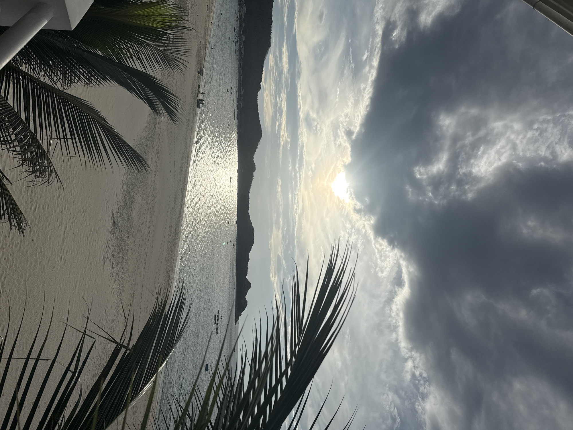 The image depicts a coastal scene at sunset in Song Cau, Vietnam, with dark, dramatic clouds partially obscuring the sun, which casts a shimmering reflection on the water. The foreground is dominated by the fronds of palm trees, framing the view of a sandy beach and the calm sea. Several small boats are scattered across the water. In the middle ground, a group of people can be seen on the beach, some walking near the water's edge and others gathered around small structures. The background features a silhouette of hills or mountains lining the coast. The overall atmosphere is serene, marked by the natural beauty of the sunset over the ocean.