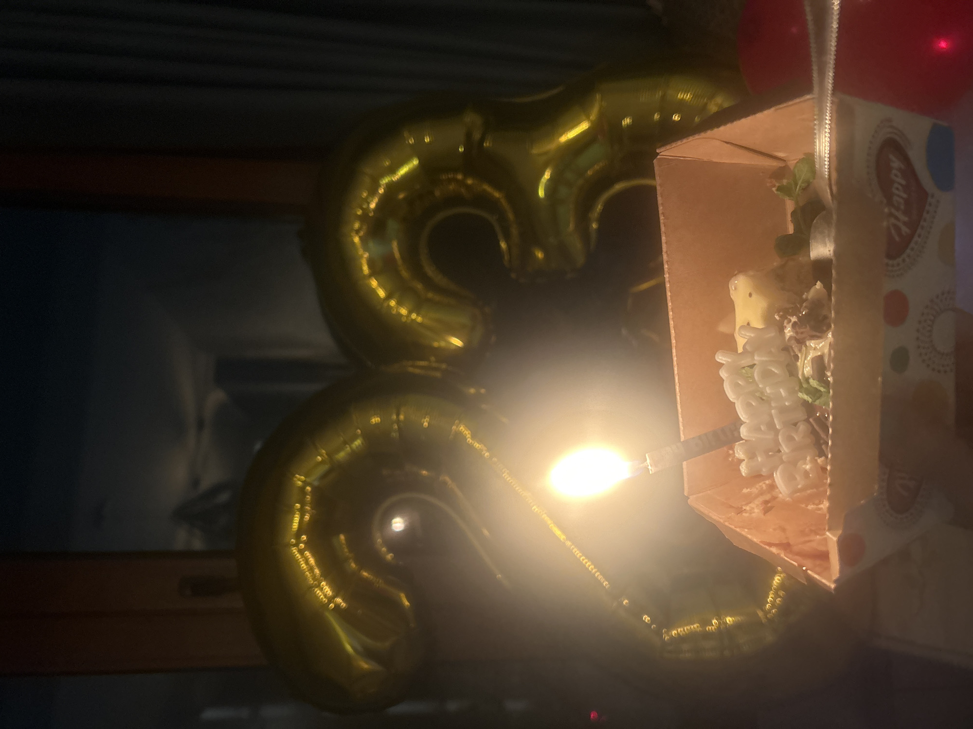 The image depicts a dimly lit indoor scene. Two gold, inflatable number balloons, resembling '33', are visible in the background. In the foreground, a birthday cake sits in a cardboard box with a lit candle inserted. The box has the word "Happy" printed on it. Two red balloons are partially visible. The location is unspecified but stated as the city of Sofia, Bulgaria.