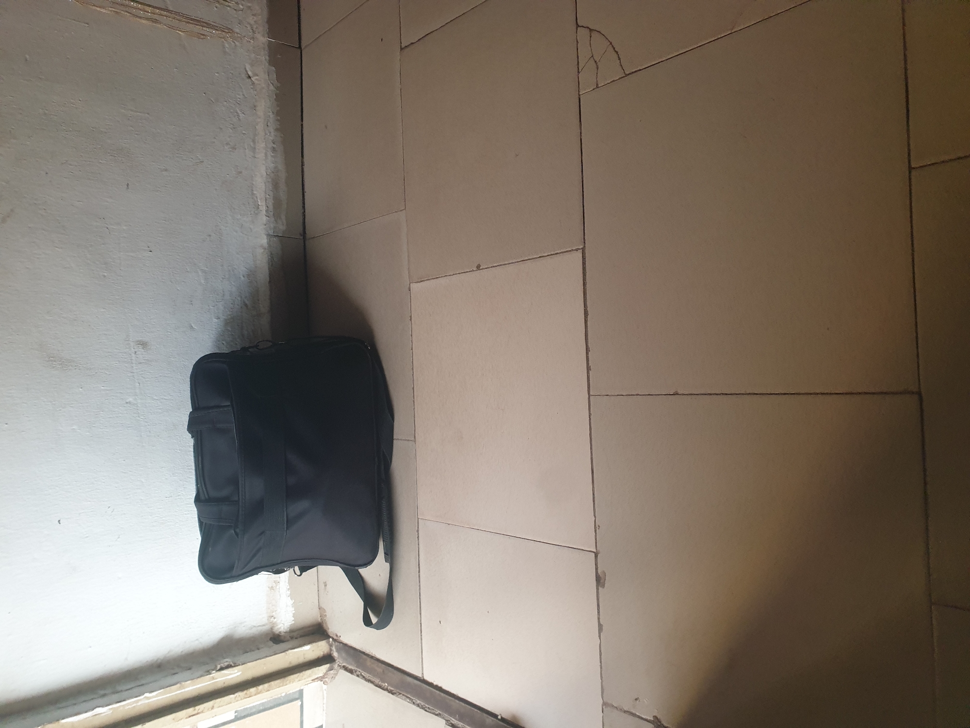 A black messenger bag rests on a tiled floor against a white wall. The bag is positioned on its side, with the strap visible. The wall appears to have some discoloration and damage near the base. The floor tiles are a light beige color and show some signs of wear and cracking. There is a doorway or opening to the left of the bag, with a metal frame. The lighting suggests an indoor environment.