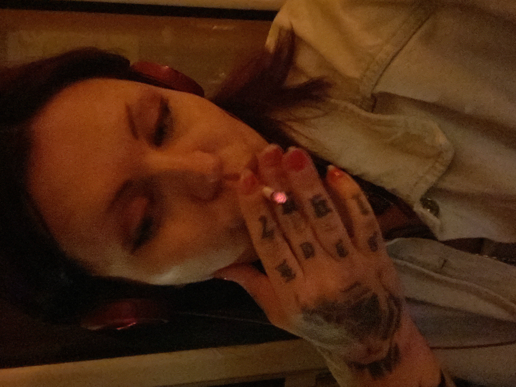A woman is lying down, holding a lit cigarette in her right hand. She has dark red hair and fair skin. Her eyes are closed. Her right hand is tattooed. She is wearing a light-colored shirt or jacket. The setting appears to be indoors. The city location is Unknown, Unknown.