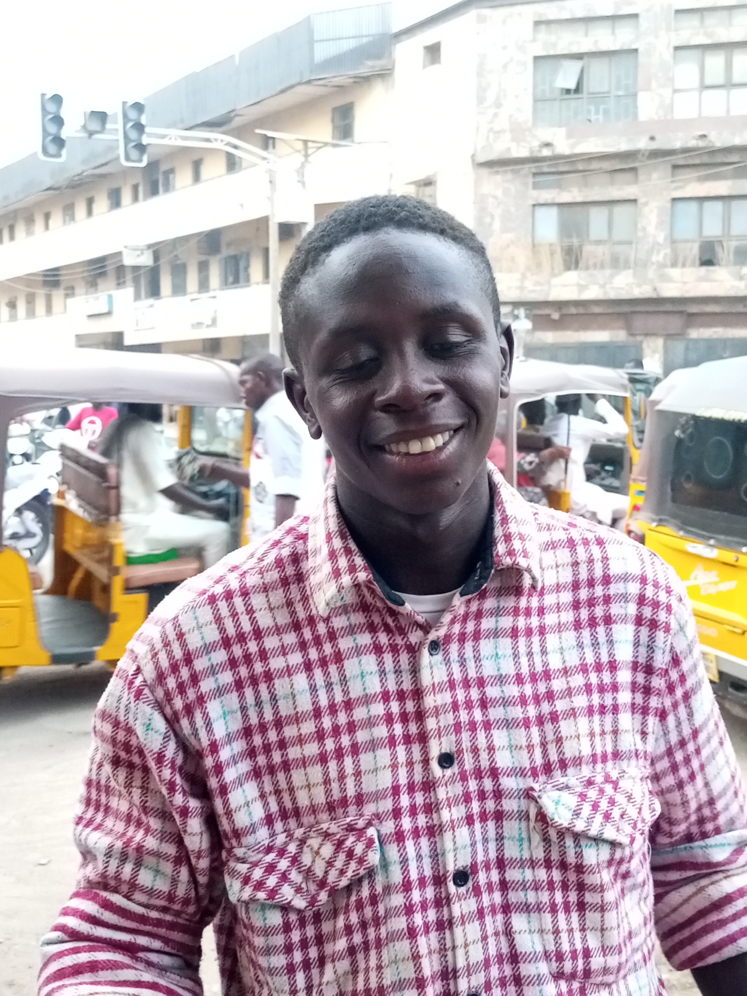 The image depicts a smiling dark-skinned man in the foreground, wearing a red and white plaid shirt with button-down pockets. Behind him are several yellow auto-rickshaws (tuk-tuks) and a building with multiple windows. A traffic light is visible on the left side of the image. The scene appears to be outdoors in a city, which is identified as Maiburiji, Nigeria. Other people are visible in the background near the tuk-tuks.