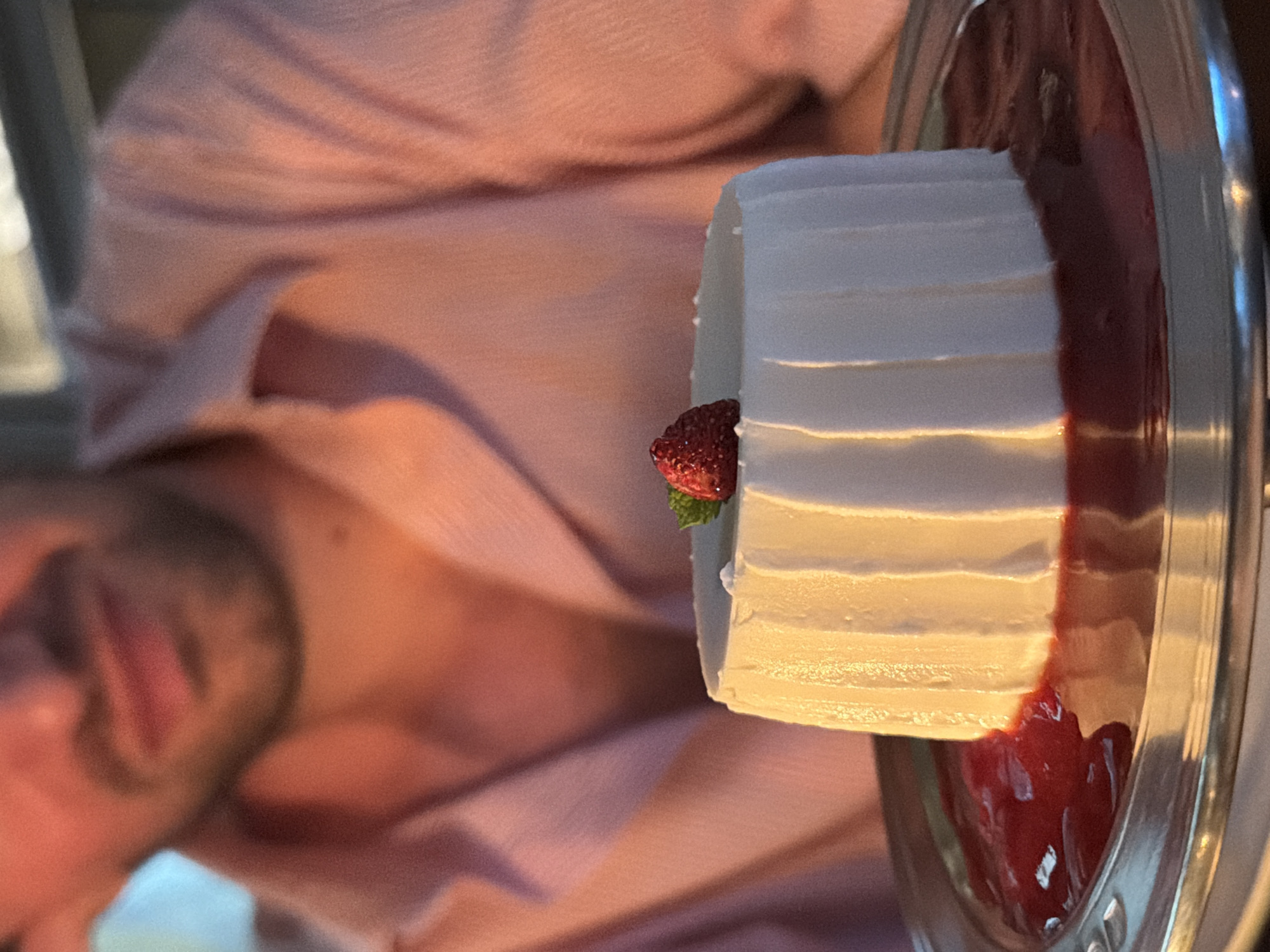 A man, partially out of focus, is seated with his head tilted downwards, looking towards a dessert. He is wearing a pink collared shirt. In the foreground, a white, cylindrical dessert is presented on a silver platter, surrounded by a red sauce. The dessert is topped with a single raspberry and a small green leaf. The lighting in the image is warm and soft, suggesting an indoor setting, possibly a restaurant. The background is blurred, but appears to be a dimly lit interior. The overall scene suggests a moment of enjoyment of a sweet treat in a sophisticated atmosphere. The image provides no specific location details about Milan, Italy.