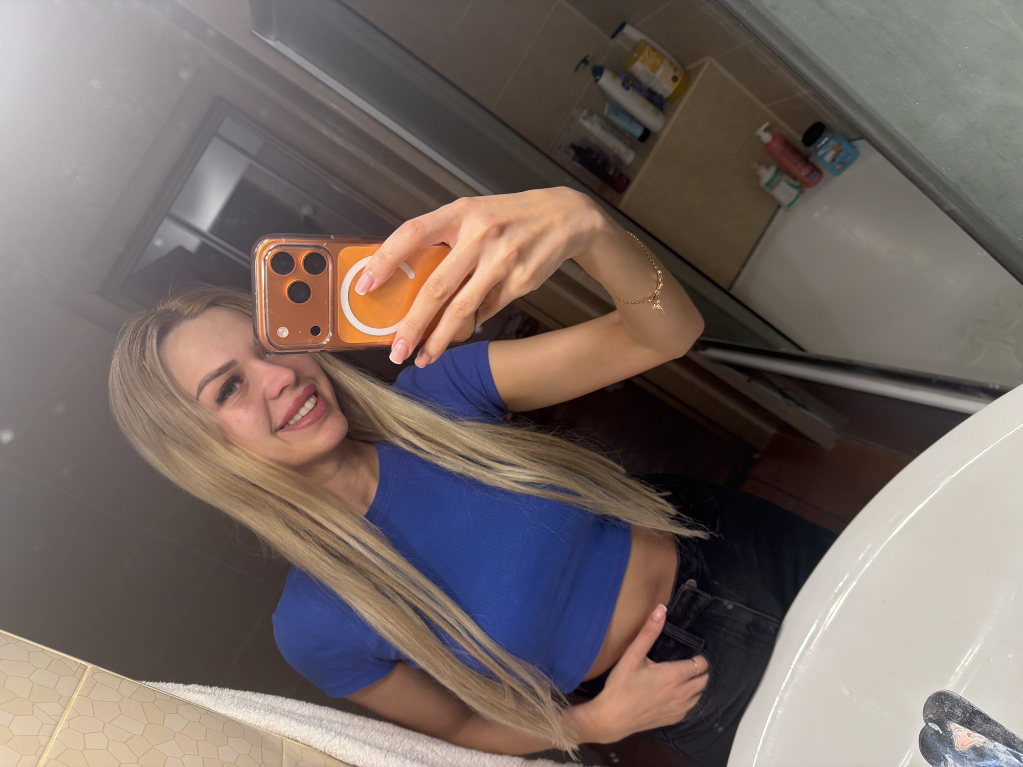 A young woman with blonde hair and a blue crop top is taking a mirror selfie in a bathroom. She is holding an orange-cased smartphone in her right hand. Her left hand is in her black jeans pocket. Behind her, on a shelf, are various toiletries. The bathroom appears to be in a residential setting.