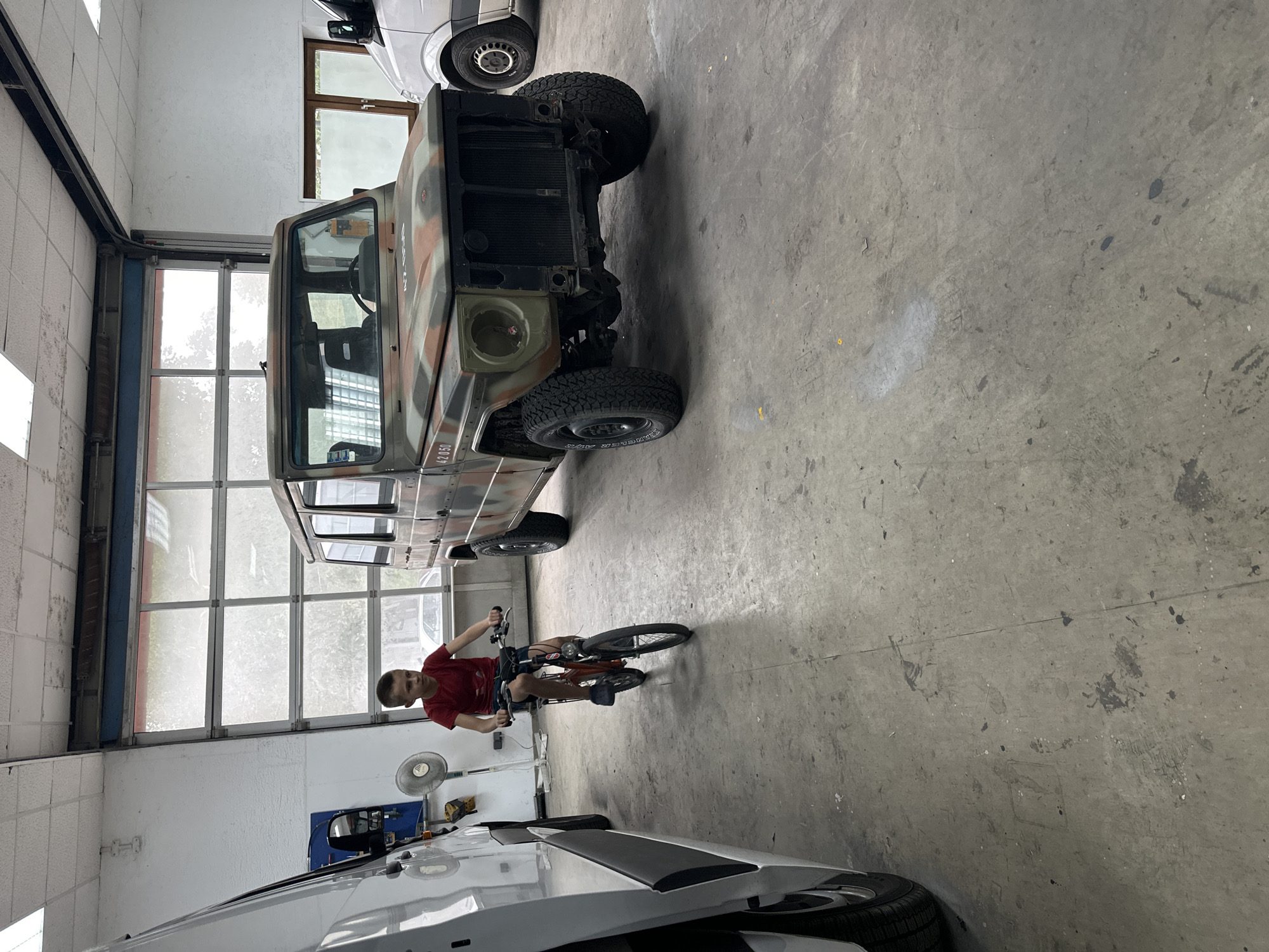 A young boy in a red t-shirt rides a small bicycle indoors, in what appears to be a garage or workshop. He is positioned in the foreground, slightly to the left of center, facing the viewer. His bicycle is an orange frame with black wheels and tires. Behind him, a large, camouflaged military-style vehicle, possibly a Mercedes-Benz G-Wagon, is parked. The vehicle's front is visible, showing a grill and headlights missing. To the left of the boy, the side of a white van is partially visible. The floor is concrete and shows signs of wear and tear, with scuff marks and stains. In the background, a large bay door with glass panels is visible, letting in natural light, suggesting it is daytime. The overall environment is utilitarian, with white walls and exposed ceiling elements. The location is identified as Mainz, Germany.