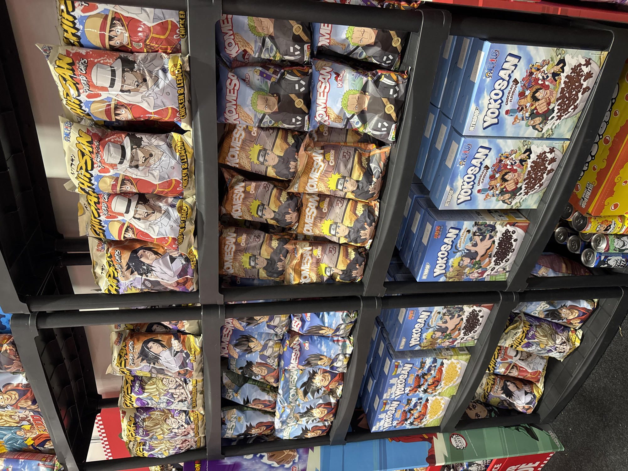 The image shows shelves in a store displaying a variety of snack food products, primarily chips and boxes of cereal, with themes from popular anime series like One Piece and Naruto. The products are arranged in plastic containers or directly on the shelves.
The subjects are the snack food products, which feature colorful packaging adorned with anime characters. These include bags of "Komesan" potato chips with various flavors like "Chicken & Lemon," "Cheese," and "Chili," as well as "Yokosan" cereal boxes labeled "Chocolate Flavour" and "Cookie Cream Flavour." There are also other snack items visible on lower shelves, some with anime imagery like Dragon Ball Z.
The scene is a retail environment, likely a supermarket or a specialty store, with the products neatly organized for sale. The lighting appears to be artificial, consistent with an indoor setting. No specific time of day or weather conditions are indicated. The overall impression is one of a well-stocked retail display aimed at fans of anime merchandise. Visible text includes brand names, product flavors, and anime titles.