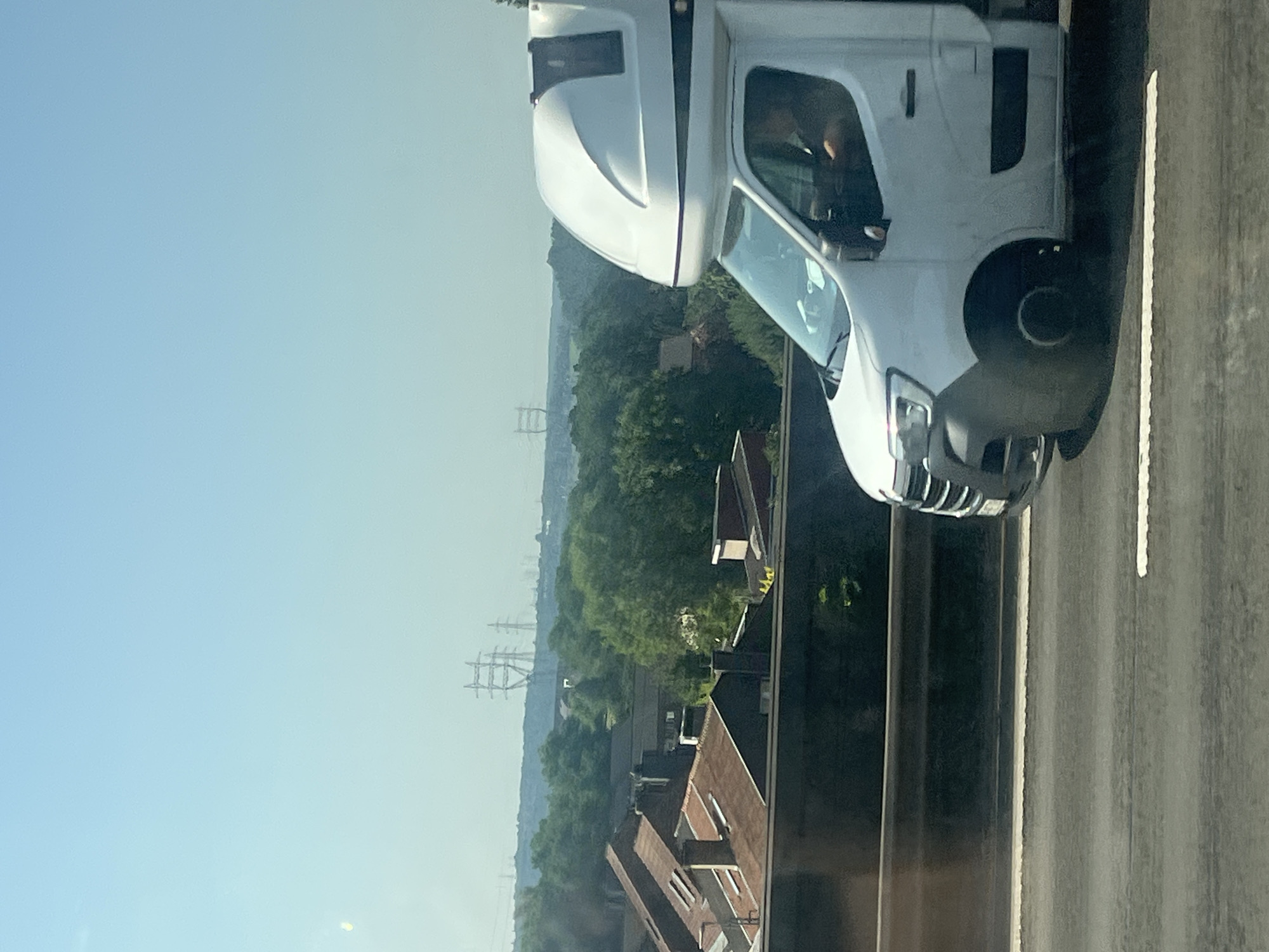 The image shows a view of the sky and a landscape from a moving vehicle. In the foreground, the side of a white van with a dark grey bumper and a raised, white cabin structure is visible, traveling on a road. The driver is partially visible in the driver's seat. The middle ground features a row of houses with reddish-brown roofs, and further beyond, a dense green forest. In the background, several tall, lattice-like electricity pylons are visible against a pale blue sky, suggesting a power grid in the distance. The lighting and hazy atmosphere indicate it is daytime, likely a sunny day. The scene appears to be a roadside view in or around Herstal, Belgium, given the presence of residential areas and significant infrastructure like power lines.
