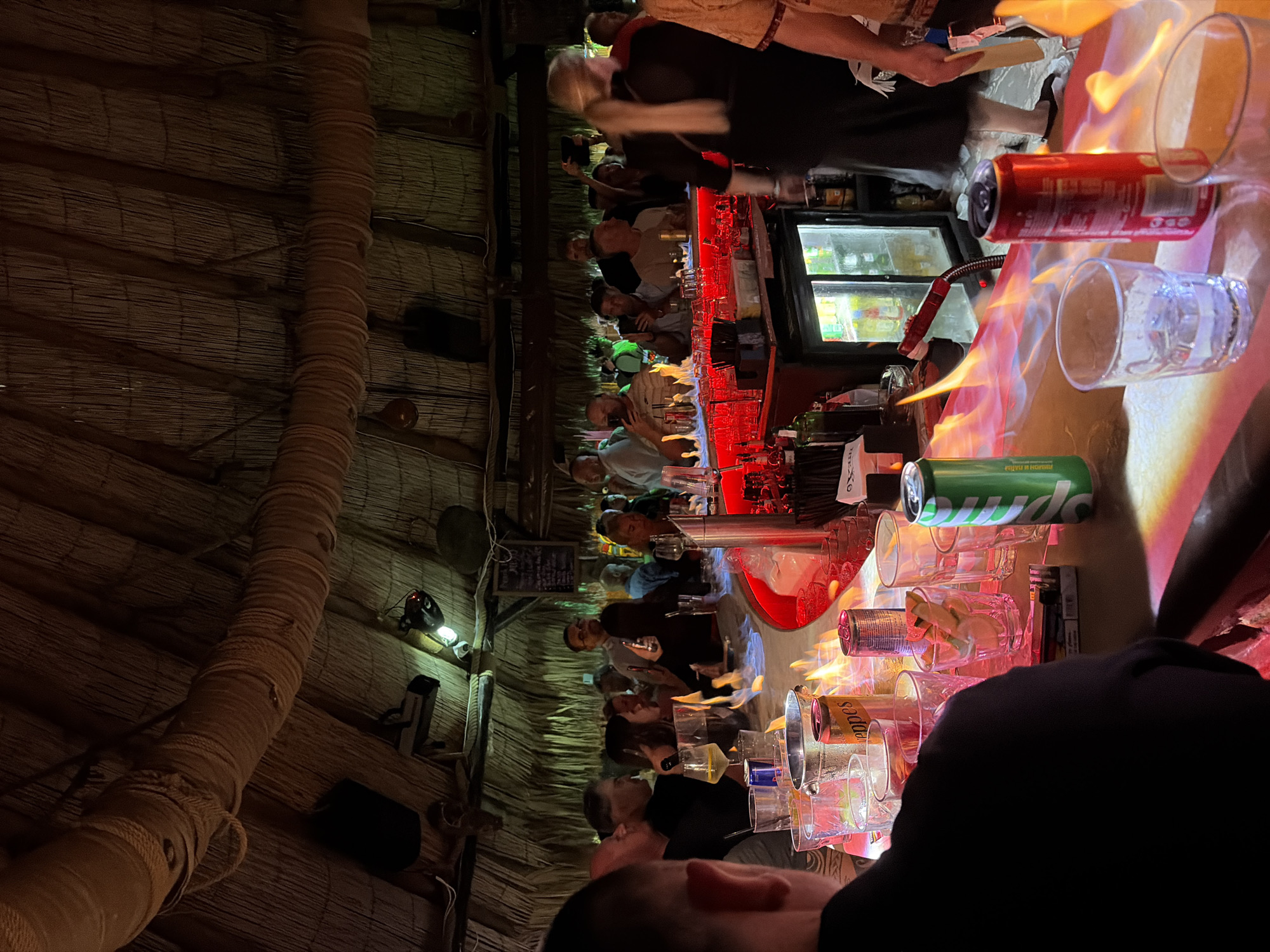 In a dimly lit, thatch-roofed bar in Nesebar, Bulgaria, a group of patrons are gathered at a counter where flames lick the surface. The bar is illuminated by red and yellow lighting, reflecting off the wet surfaces of glasses and cans. People are standing and sitting, some are holding drinks and phones, and the overall atmosphere appears lively and social. A bartender, partially visible, stands behind the counter, and patrons are engaged in conversations. The bar is decorated with ropes and has a rustic, tropical feel.