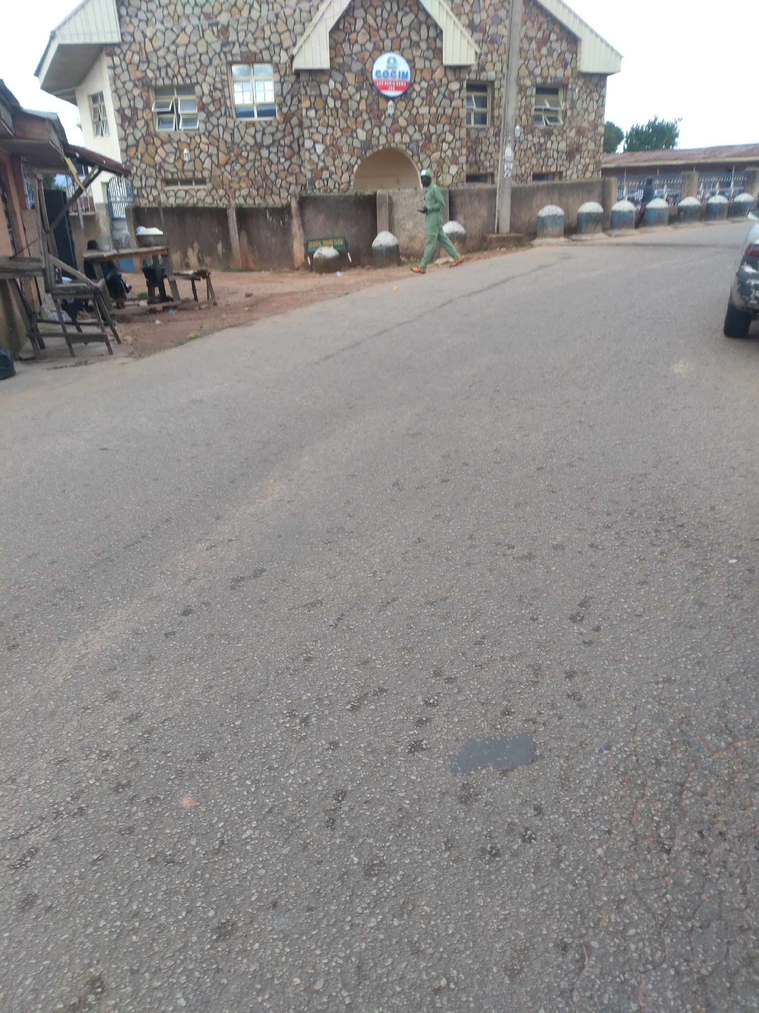 A full outdoor shot shows a building with a stone facade and a red and blue sign that says "COCIN CHURCH". A person in a green uniform is walking on the paved road in front of the building. The building is located in Jos, Nigeria. The road is gray and cracked. There are several round, white-topped bollards lining the side of the road. In the foreground on the right, a portion of a car is visible. The background shows more buildings and trees.