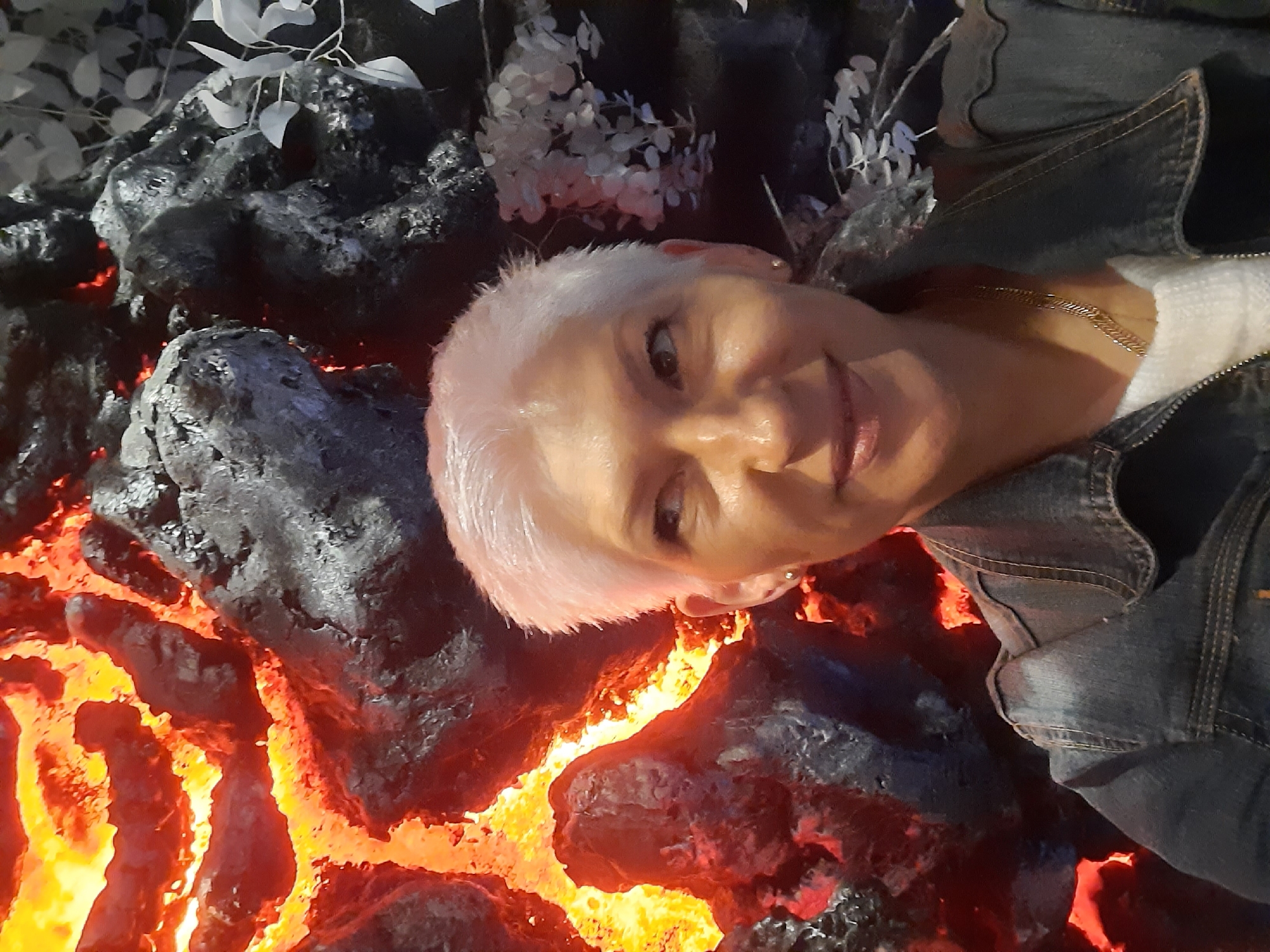 A close-up, selfie-style photo shows a fair-skinned woman with short, white hair and subtle makeup, looking to her left. She is wearing a denim jacket over a white top and a gold necklace. Behind her, a backdrop features what appears to be a stylized representation of lava, with bright orange and red glowing fissures contrasted against dark, rocky textures. To the right, a decorative element resembling pale leaves is visible. The overall impression is of an indoor setting, possibly an exhibit or a themed space. The location is identified as Minsk, Belarus.