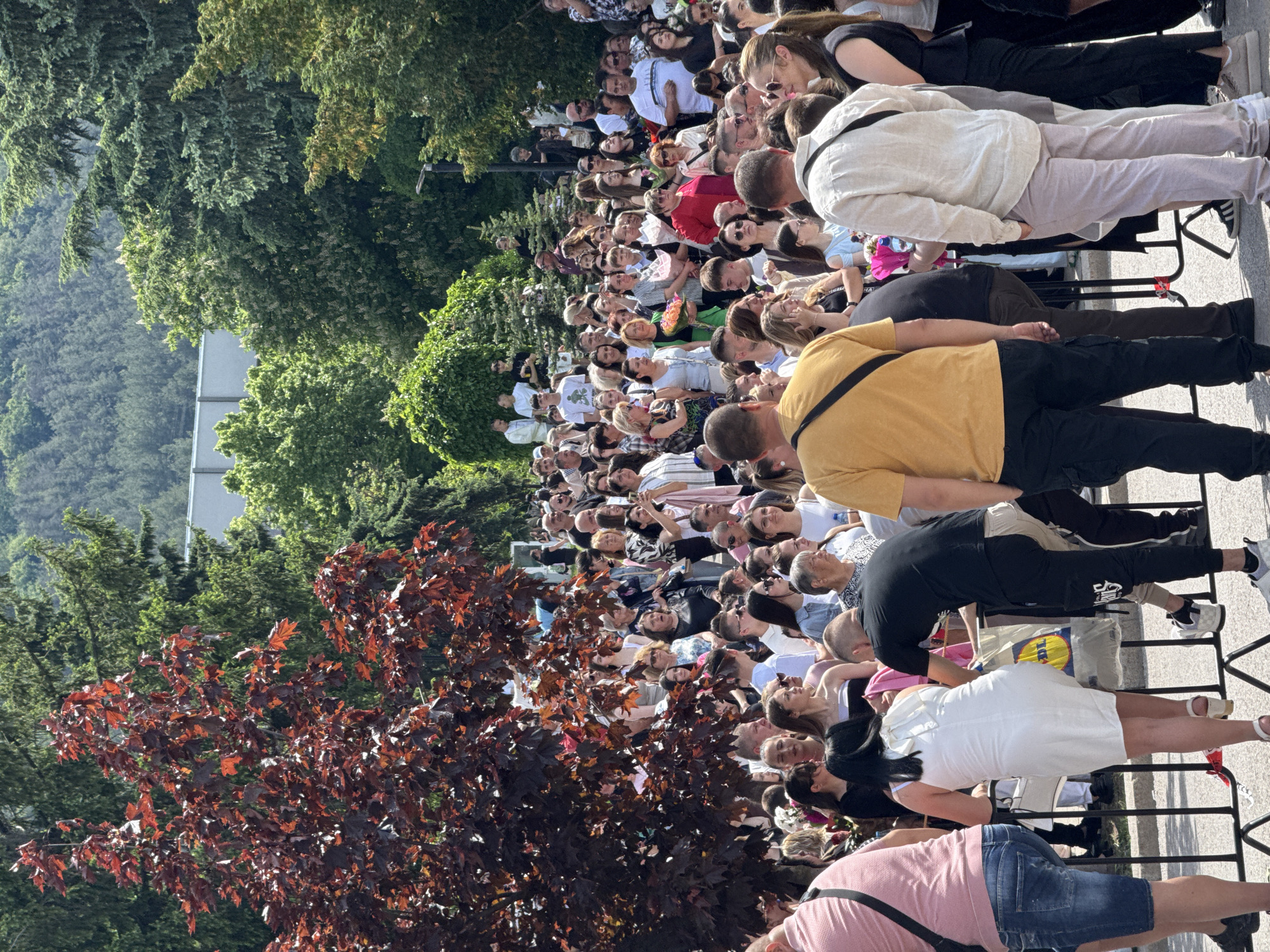 The image depicts a large crowd of people gathered outdoors, possibly attending an event or public gathering. The crowd is dense, with individuals of various ages and appearances. Many are looking in the same direction, suggesting a focal point of interest. Trees and a building are visible in the background, indicating an urban or suburban setting. The location is Shumen, Bulgaria, suggested by the context. Metal crowd-control barriers are present, likely to manage the crowd. Some individuals are wearing casual attire, while others are dressed more formally. The overall atmosphere seems expectant and attentive.