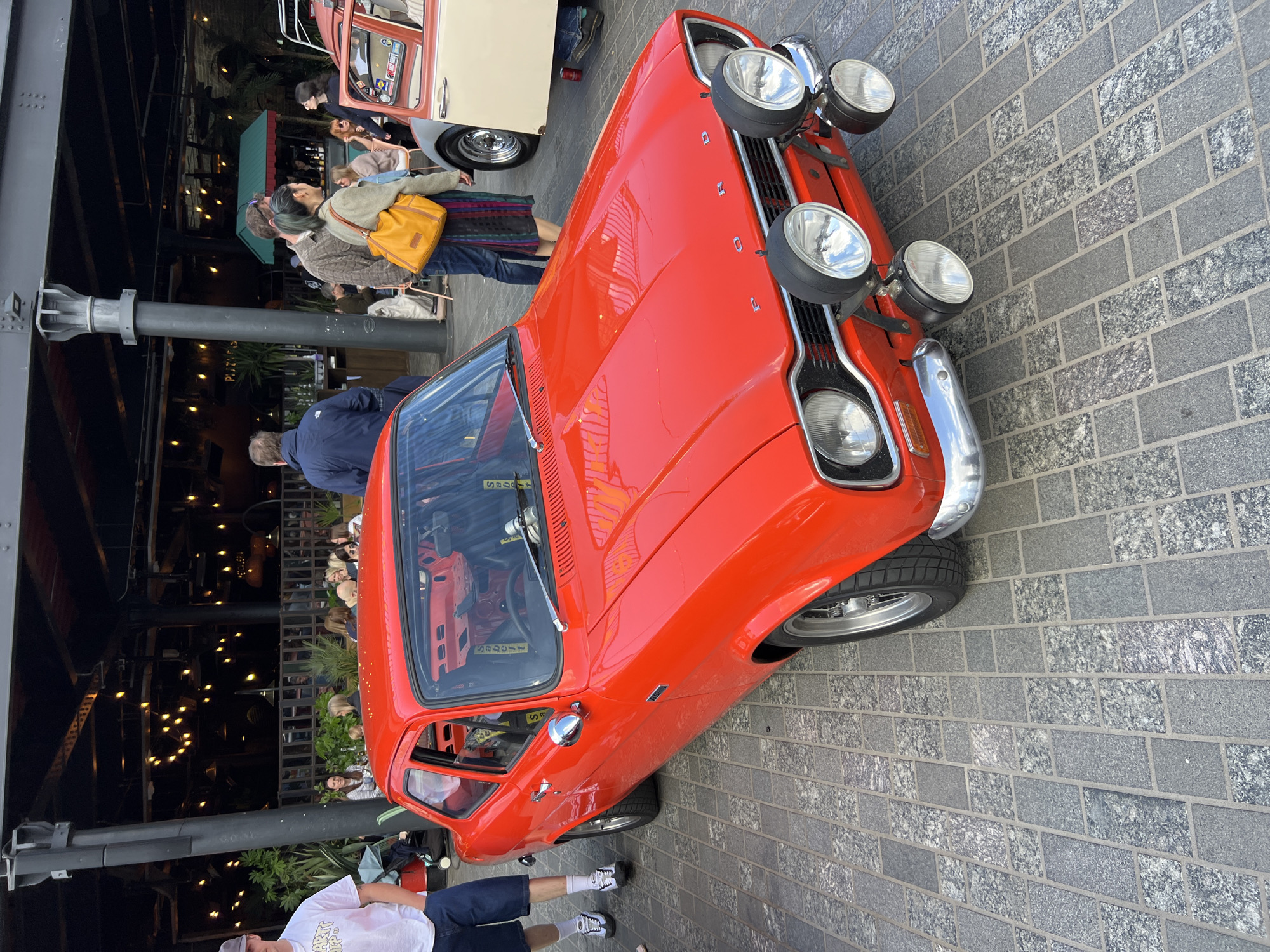 The image shows a red Ford Escort Mark I parked on a grey brick-paved area. The car has four auxiliary lights mounted on the front. People are standing around the car and in the background. There are buildings and light fixtures visible behind the car. The location appears to be a public area, possibly near a restaurant or bar. The setting and presence of vintage cars suggest it might be a car show or gathering in an urban environment, potentially in London, United Kingdom, based on the car's British origins and the architectural style.