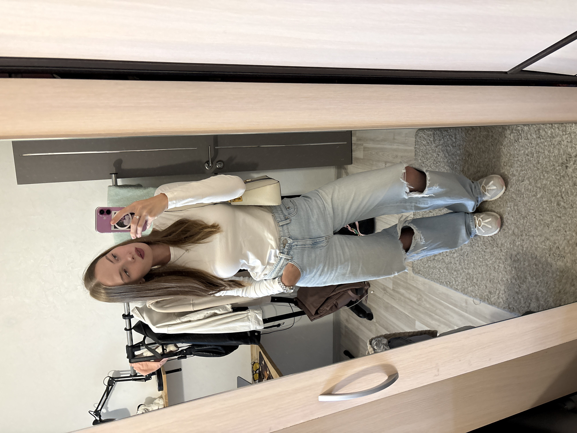 A young woman is taking a mirror selfie in what appears to be a bedroom or dressing area. She is wearing a white long-sleeved shirt and light-wash ripped jeans. She is also wearing white sneakers and carrying a small, light-colored purse over her shoulder. The background shows a closet or wardrobe area with clothes hanging and a mirror reflecting the scene. The overall lighting suggests it could be daytime indoors. The woman has long brown hair and is looking at her phone, which is held up to take the picture. She is wearing makeup, with lipstick visible. A ring is on her left hand. The floor is carpeted in a neutral color and there are wooden furniture elements visible, including a drawer with a silver handle. The image is oriented vertically, as if taken with a phone held in portrait mode.