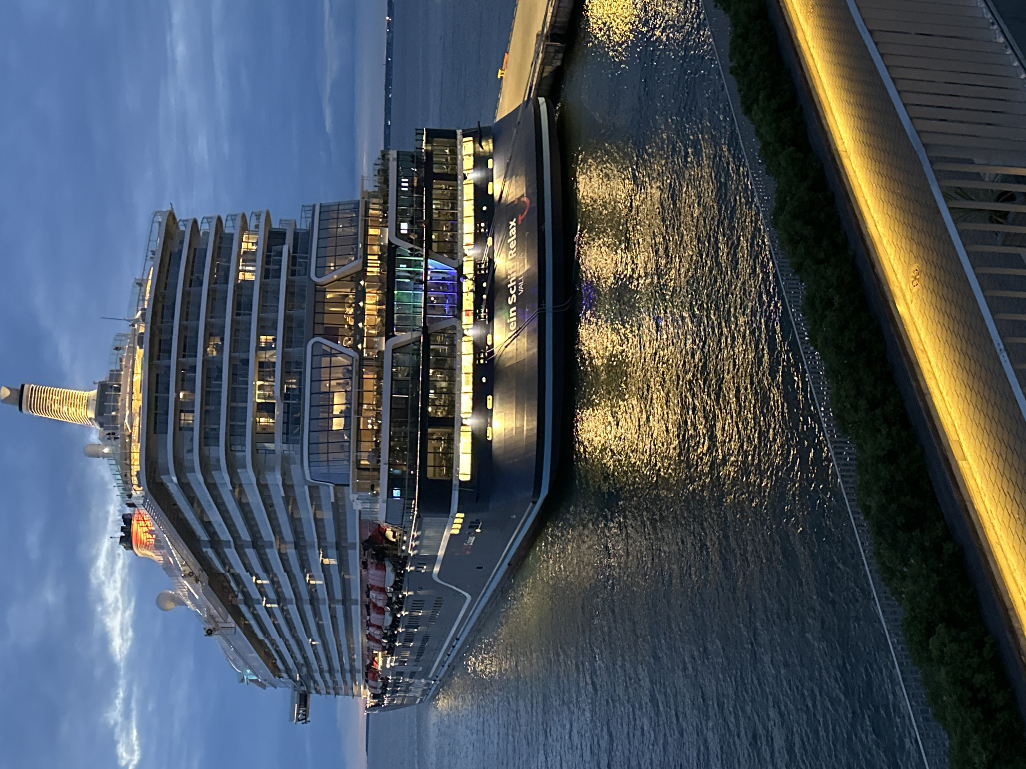 A large cruise ship, the "Mein Schiff Relax," is docked at a pier in Tallinn, Estonia. The ship is illuminated by numerous lights, casting a warm glow on the water. The sky is a deep blue, indicating it is either dusk or dawn. The ship is dark blue with white railings and decks. There are multiple lifeboats visible on the side of the ship. The pier is made of concrete, and there is a railing along the edge. In the foreground, there is a walkway with yellow lights and some greenery. The overall scene is calm and serene, with the ship appearing majestic against the twilight sky.
