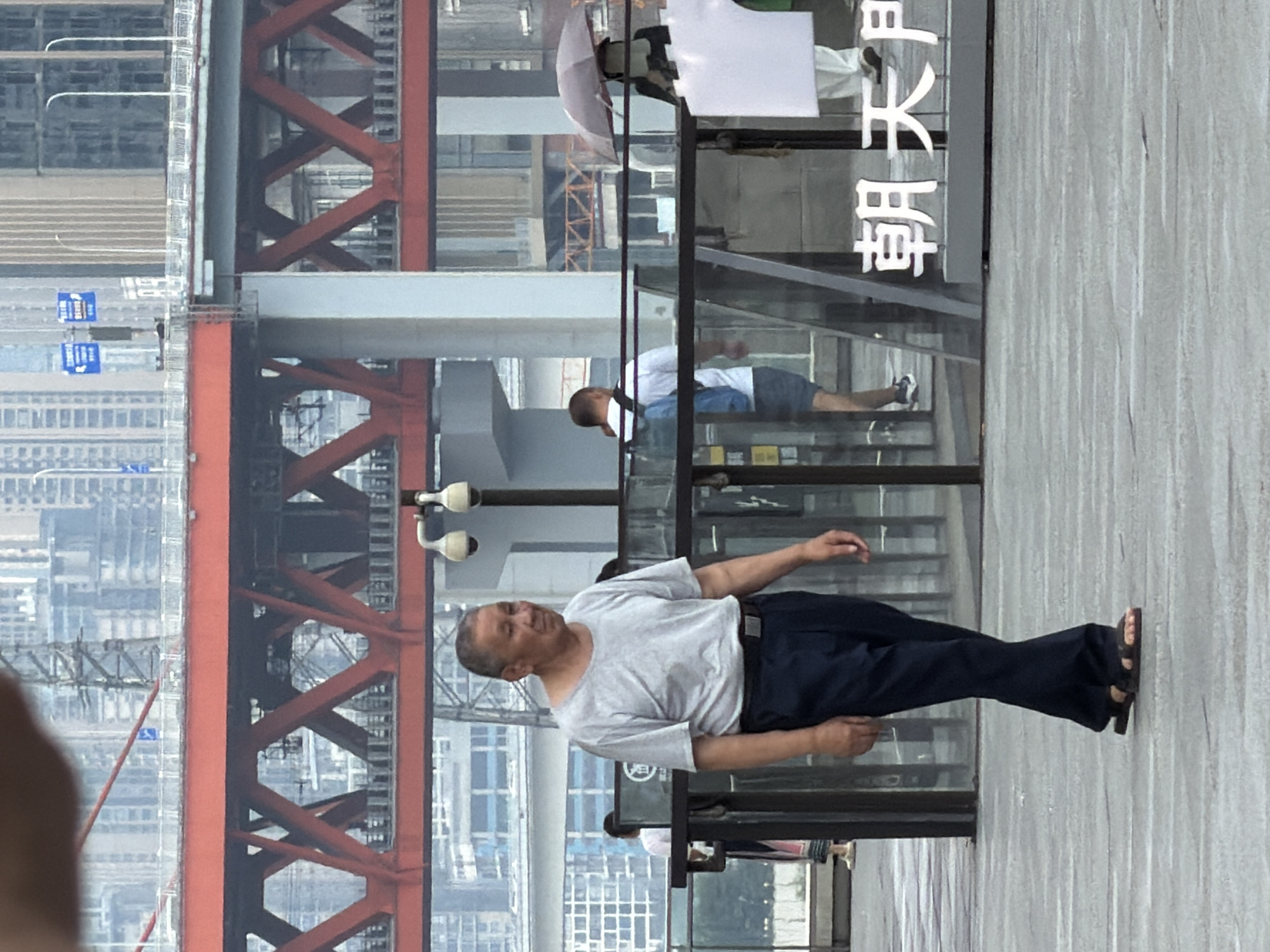 An elderly East Asian man walks forward in the foreground, wearing a grey t-shirt, dark pants, and sandals. He is on a paved area with visible white lines. Behind him, a transparent barrier encloses a walkway with glass panels. Beyond the barrier, other people are visible, including a man with a backpack and a person holding an umbrella. In the background, a large red bridge spans across the scene, with numerous tall buildings in the distance. The environment appears to be an urban area in Yuzhong District, China.