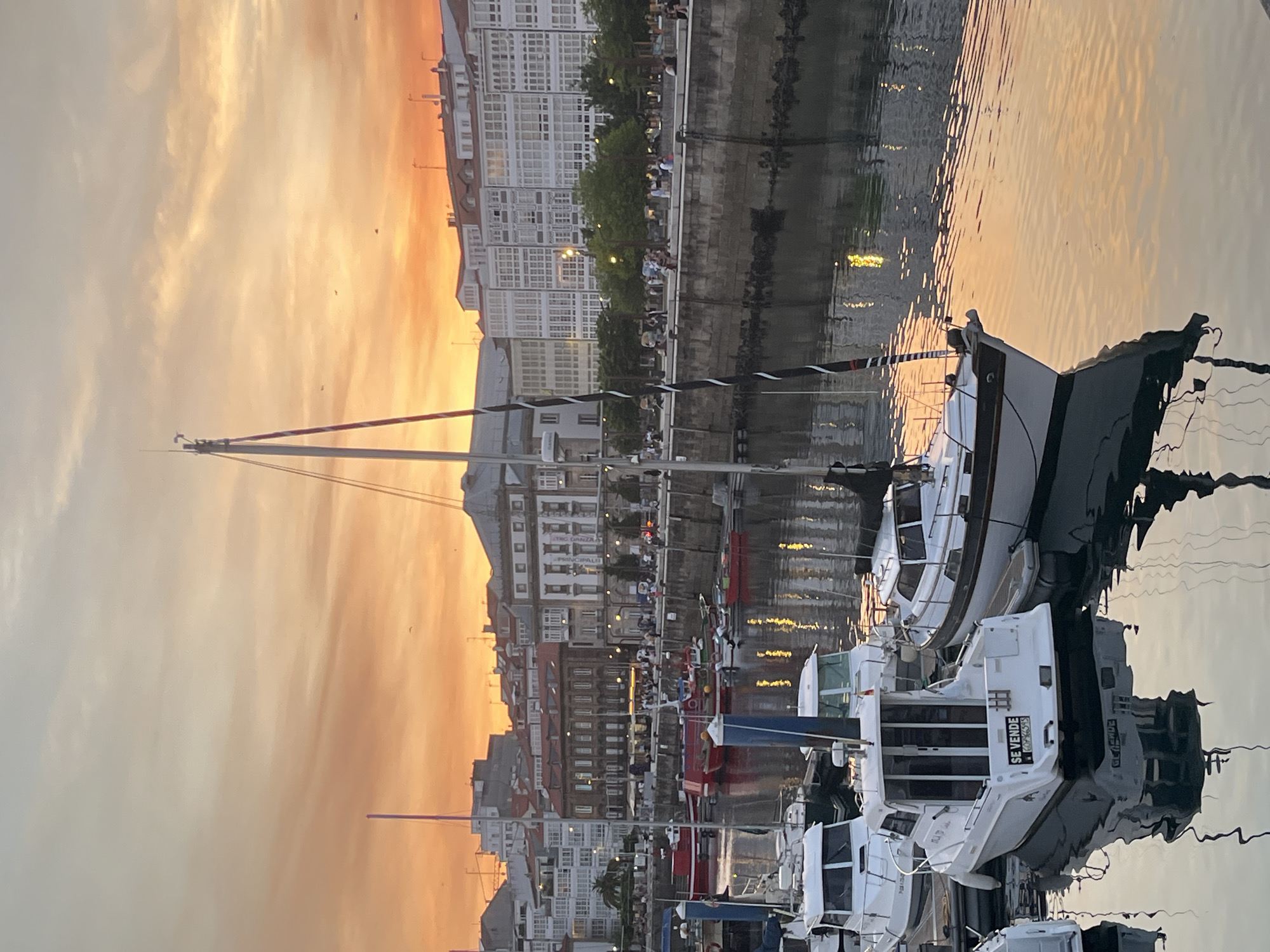 The image depicts a harbor in A Coruña, Spain, at sunset. The sky is a vibrant orange and yellow, with some clouds. In the foreground, several boats are docked. One white motorboat in the center has "SE VENDE" (For Sale) written on its stern. Other boats, including sailboats and smaller vessels, are also visible. Behind the boats, a stone quay lines the waterfront, with a walkway on top where numerous people are strolling. Beyond the quay, a row of buildings with white balconies faces the harbor. The water reflects the colorful sky and the lights from the buildings. The overall scene conveys a peaceful evening atmosphere in a bustling port city.