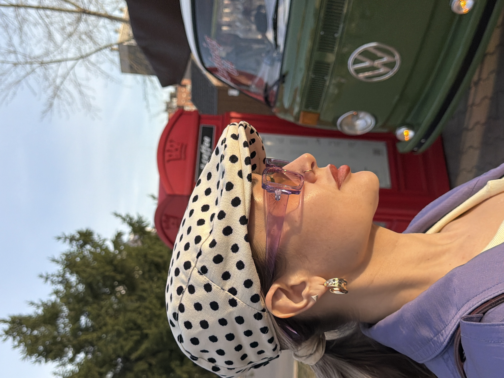 A woman wears a white and black polka dot hat and purple glasses and a purple jacket in a low-angle outdoor shot. She is positioned in front of a red phone booth, with a green Volkswagen bus parked behind. The scene is outdoors, indicated by a tree with bare branches and a blue sky in the background. A building is partially visible behind the phone booth. The location is Krasnoyarsk, Russia, evidenced by the red telephone box resembling the traditional British phone booth.