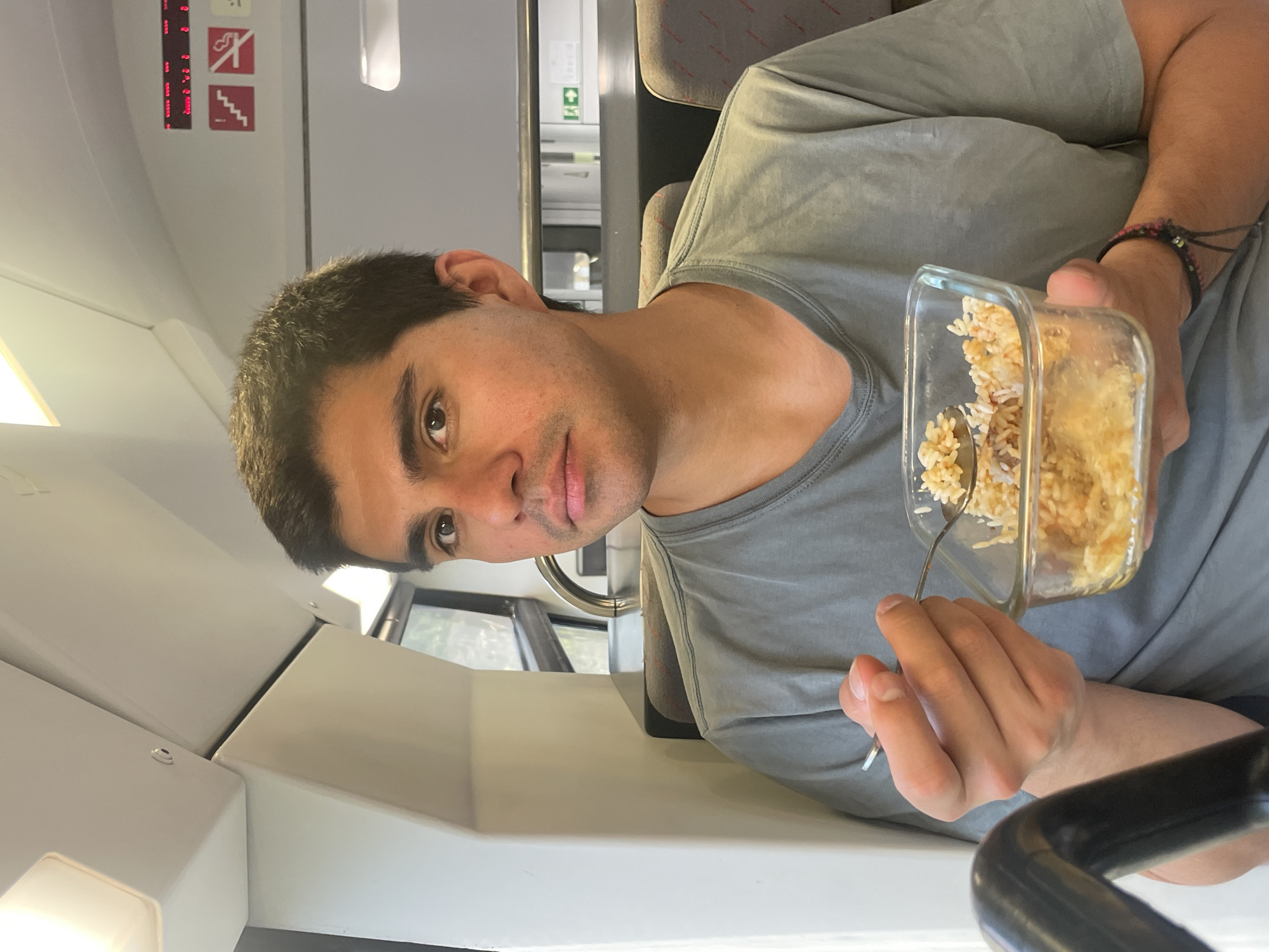 A young man with short, dark hair is seated on a train, holding a clear glass container filled with rice and a spoon. He is looking towards the camera with a neutral expression. The train interior features grey seats with red accents, a window with a view of greenery, and overhead compartments. Safety signs, including one depicting stairs and another prohibiting smoking, are visible. The scene appears to be in Gavà, Spain, suggested by the train context.