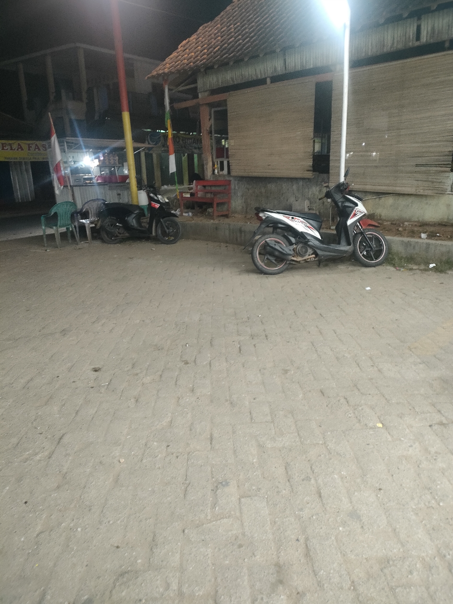 A nighttime street scene in Desa Banyumas, Indonesia, depicts two parked motorcycles on a paved walkway. The motorcycle on the left is black, and the one on the right is white with red and black accents. In the background, a building with a tiled roof and bamboo-like siding is visible, along with a streetlamp that illuminates the area. Red chairs and a table are also present near the building. Flags of Indonesia, white and red, are displayed on poles near the building entrance. The ground is covered with paving stones, and some debris is scattered around.